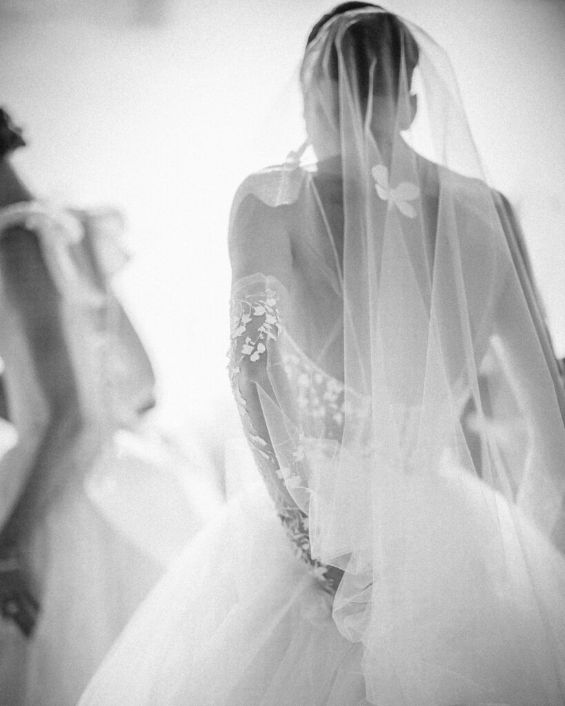 A bride is captured from the back in a stunning black and white image, her wedding dress and veil adorned with intricate lace details. The elegance of the Pnina Tornai AURA Collection shines through in this timeless moment.