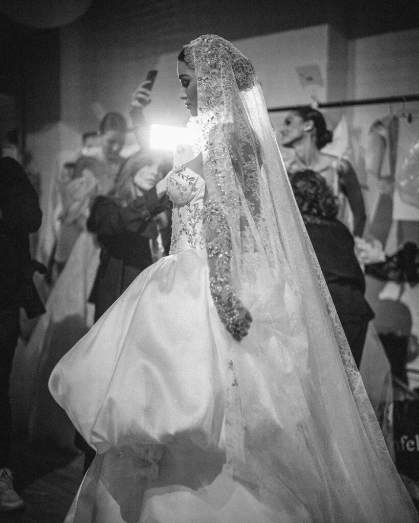 A bride in a long, lace veil and an elegant gown from the Pnina Tornai AURA Collection stands amidst others backstage, enveloped by activity.