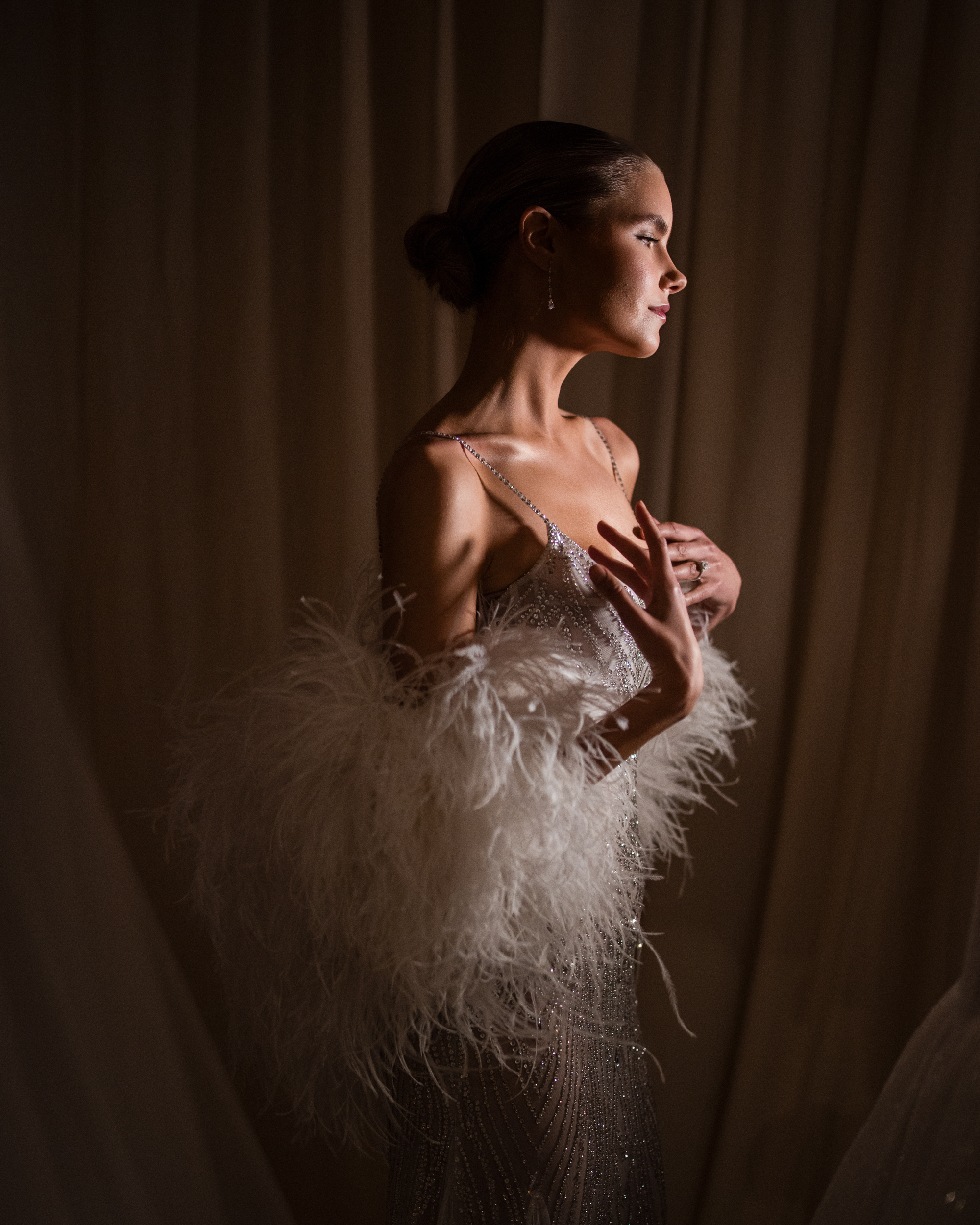 A woman in an embellished dress from the Pnina Tornai AURA Collection, featuring intricate feather details, stands in profile against a draped curtain, softly lit from the side.