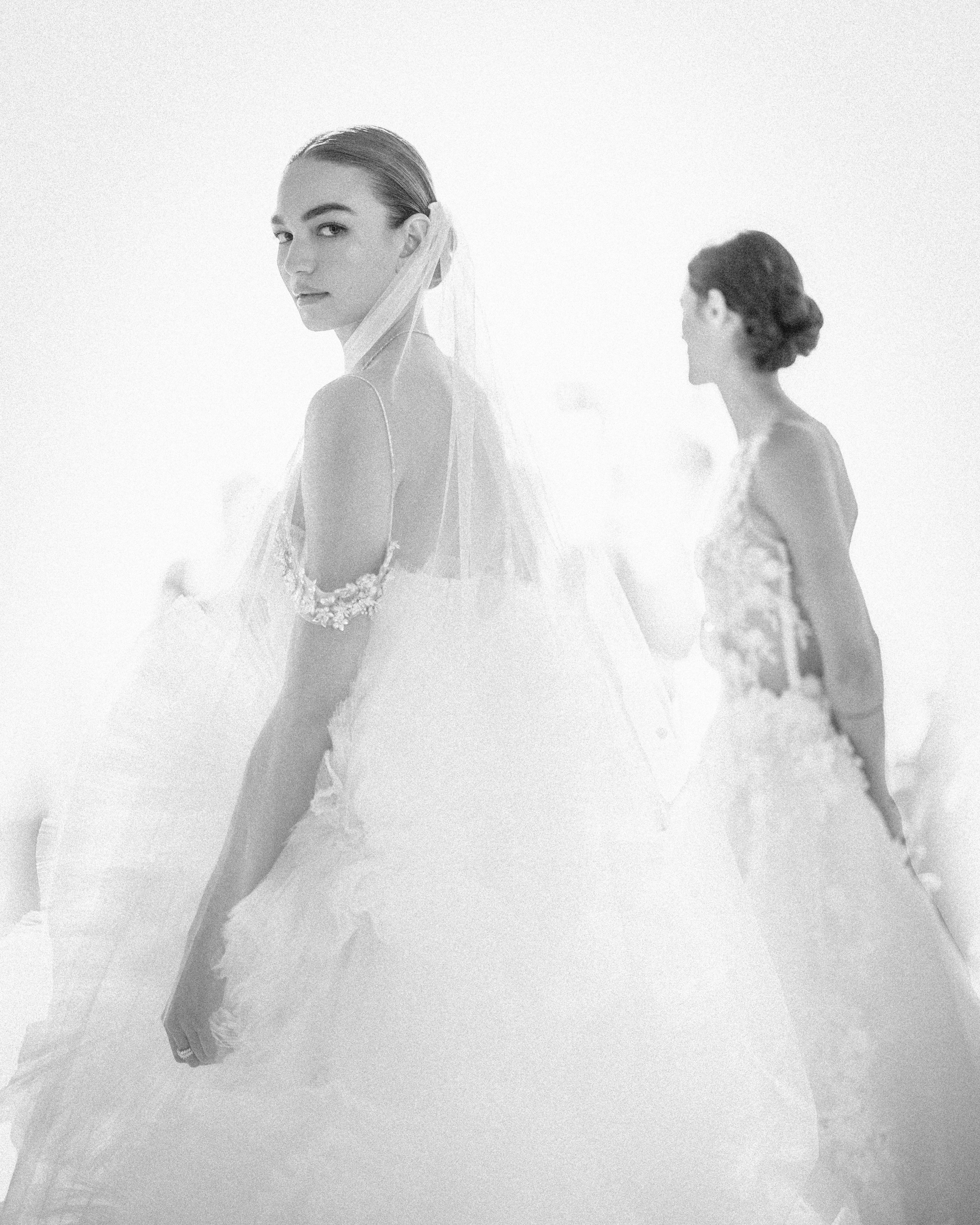 Two women in exquisite bridal gowns from the Pnina Tornai AURA Collection; one faces forward, the other turns away. The setting is bright and ethereal, enhancing the intricate details of their attire.