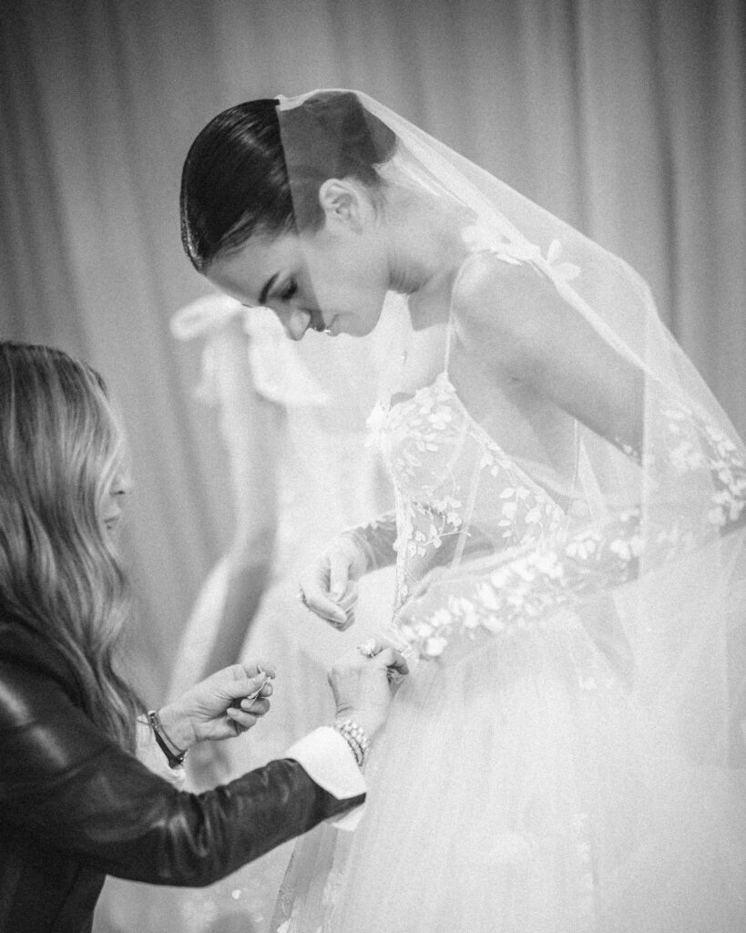 A woman delicately adjusts a bride's wedding dress train in a softly lit room. The bride, wearing an elegant veil and an embroidered gown from the Pnina Tornai AURA Collection, stands beautifully poised for her big moment.