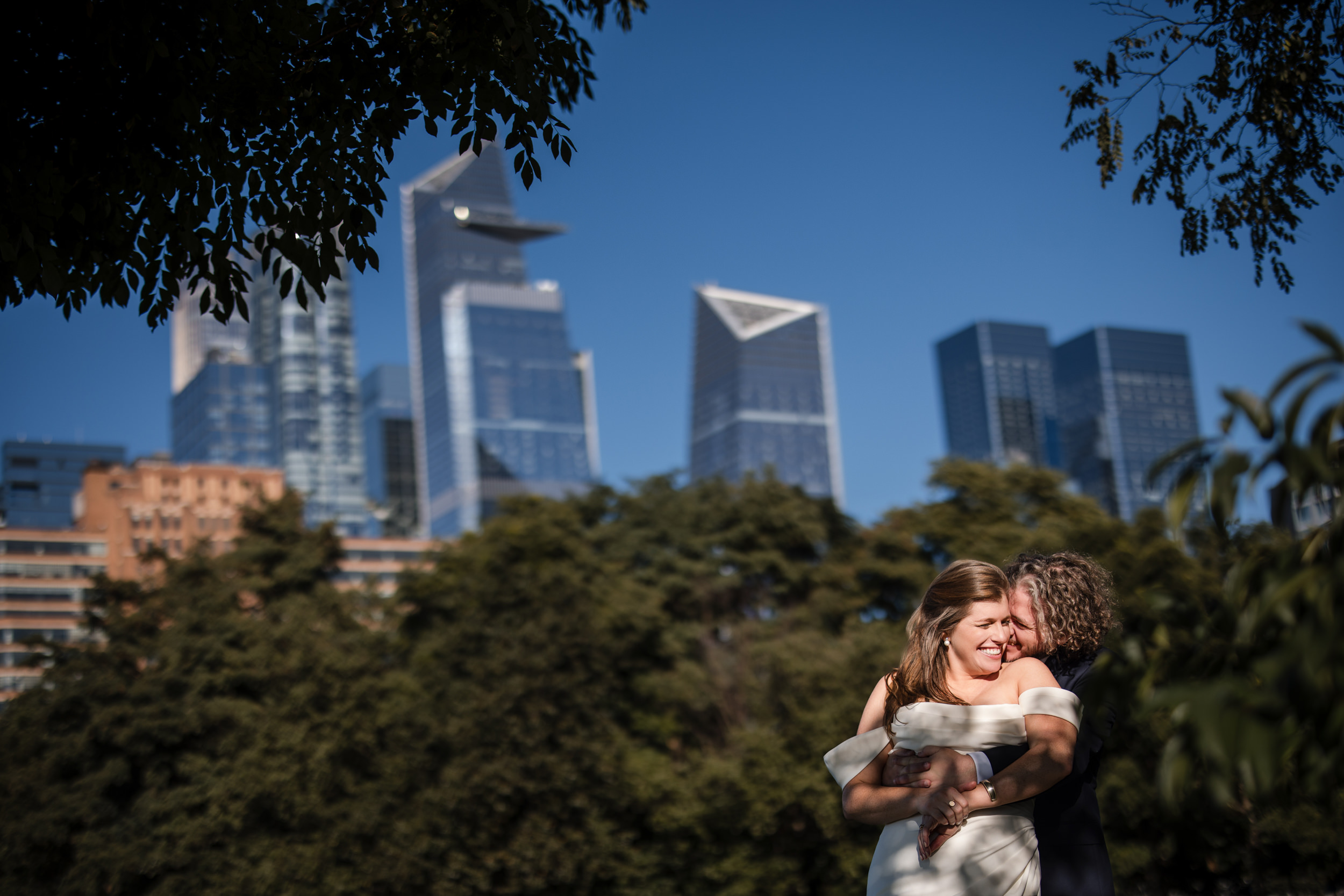 In the leafy park, two people embrace with a city skyline behind them, savoring a moment reminiscent of a romantic Greenwich Hotel wedding on a clear day.