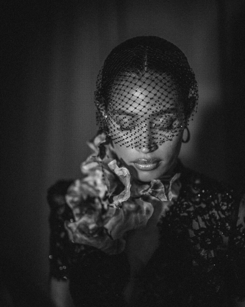 A black and white image captures the essence of the Pnina Tornai AURA Collection, featuring a person wearing a netted veil and floral accessory. Their eyes are gently closed under dramatic lighting, evoking elegance and mystery.