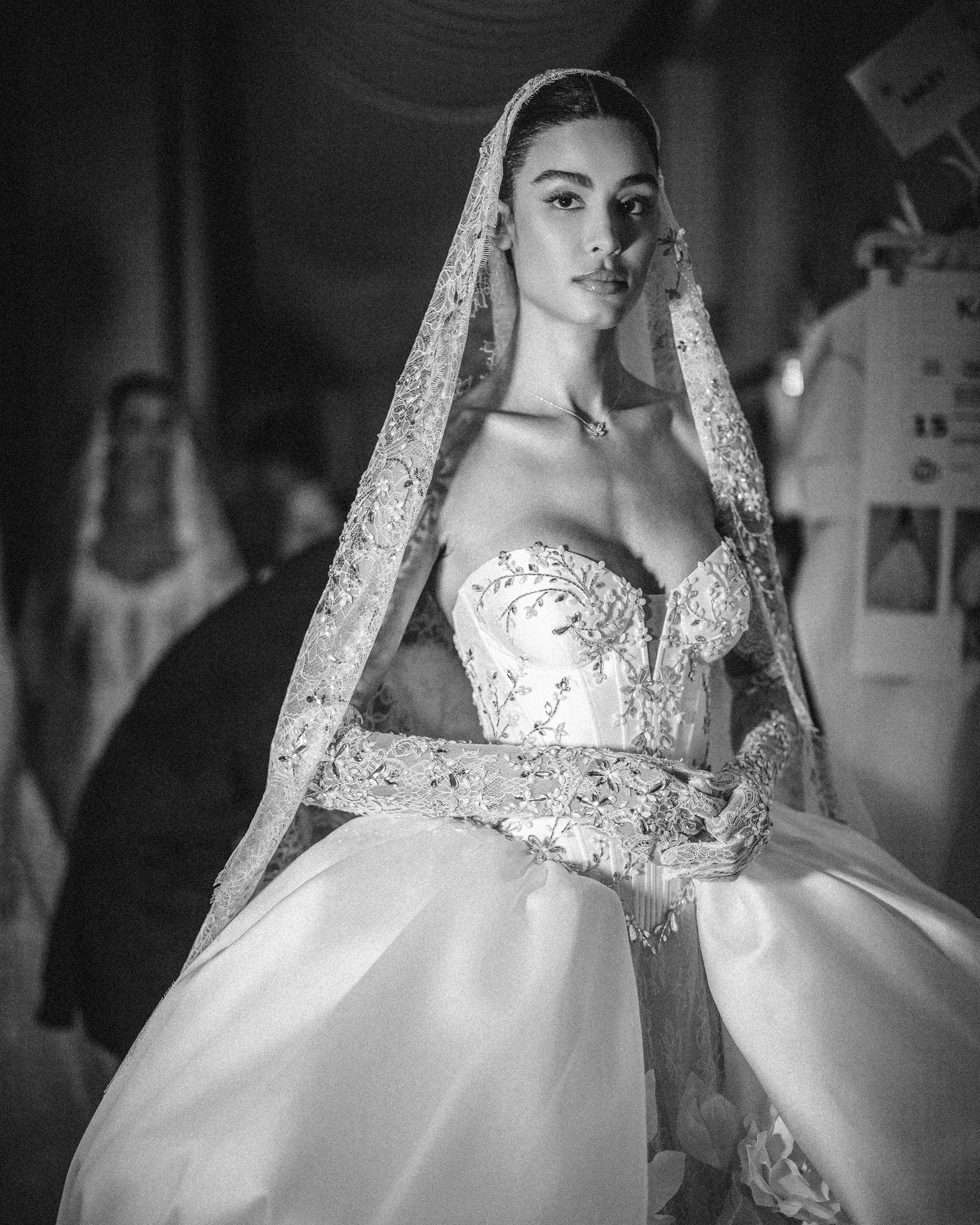 A model in an intricately detailed wedding gown from the Pnina Tornai AURA Collection poses backstage, her long veil cascading gracefully. The dress, captured in black and white, features exquisite floral embroidery and voluminous sleeves.