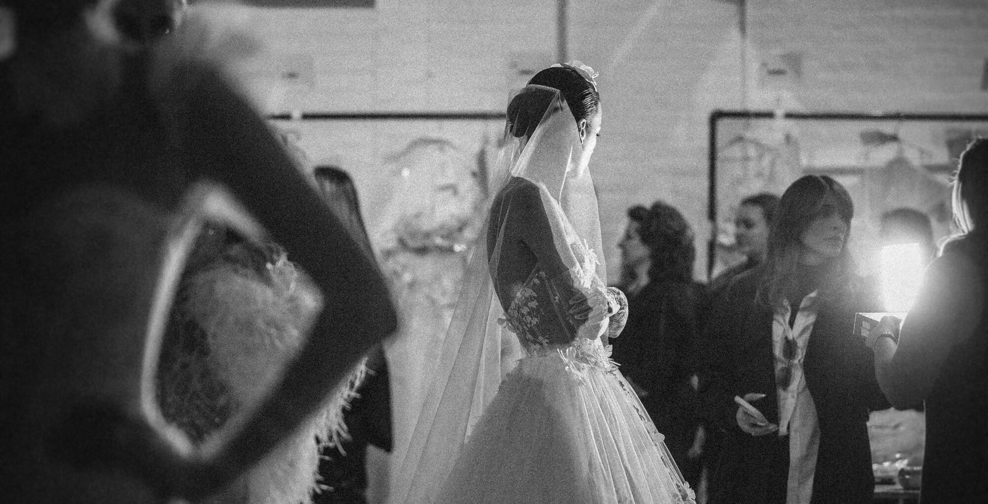 A bride in a Pnina Tornai gown from the AURA Collection stands gracefully among people in a dimly lit room.