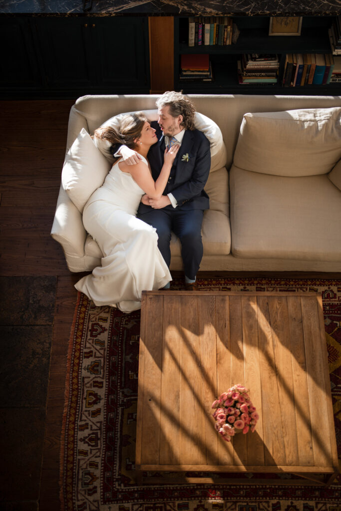 A couple in formal wedding attire lies on a sofa, embracing warmly at the elegant Greenwich Hotel. In front of them, a wooden table with a pink bouquet rests gracefully on a patterned rug.