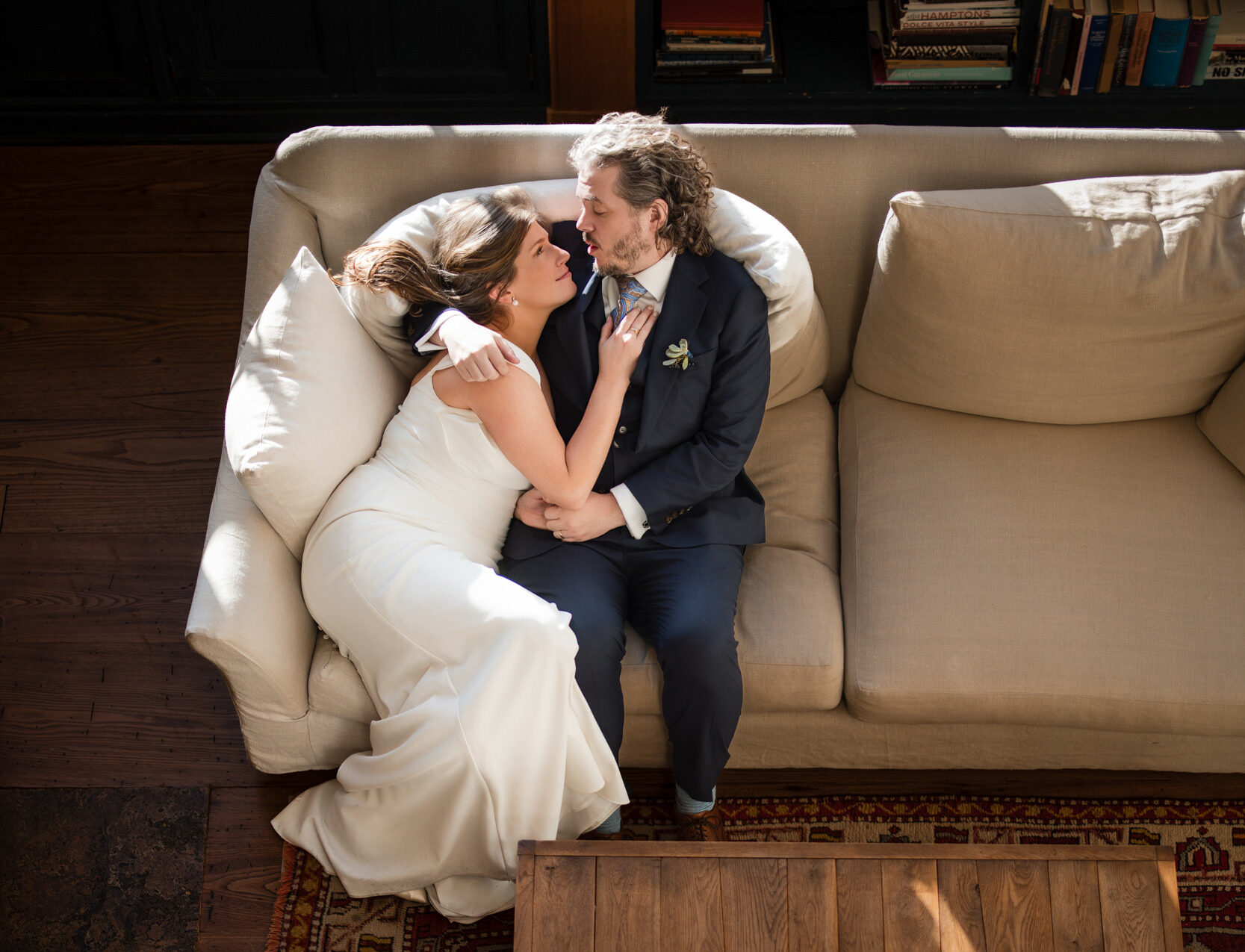 A couple in formal wedding attire lies on a sofa, embracing warmly at the elegant Greenwich Hotel. In front of them, a wooden table with a pink bouquet rests gracefully on a patterned rug.