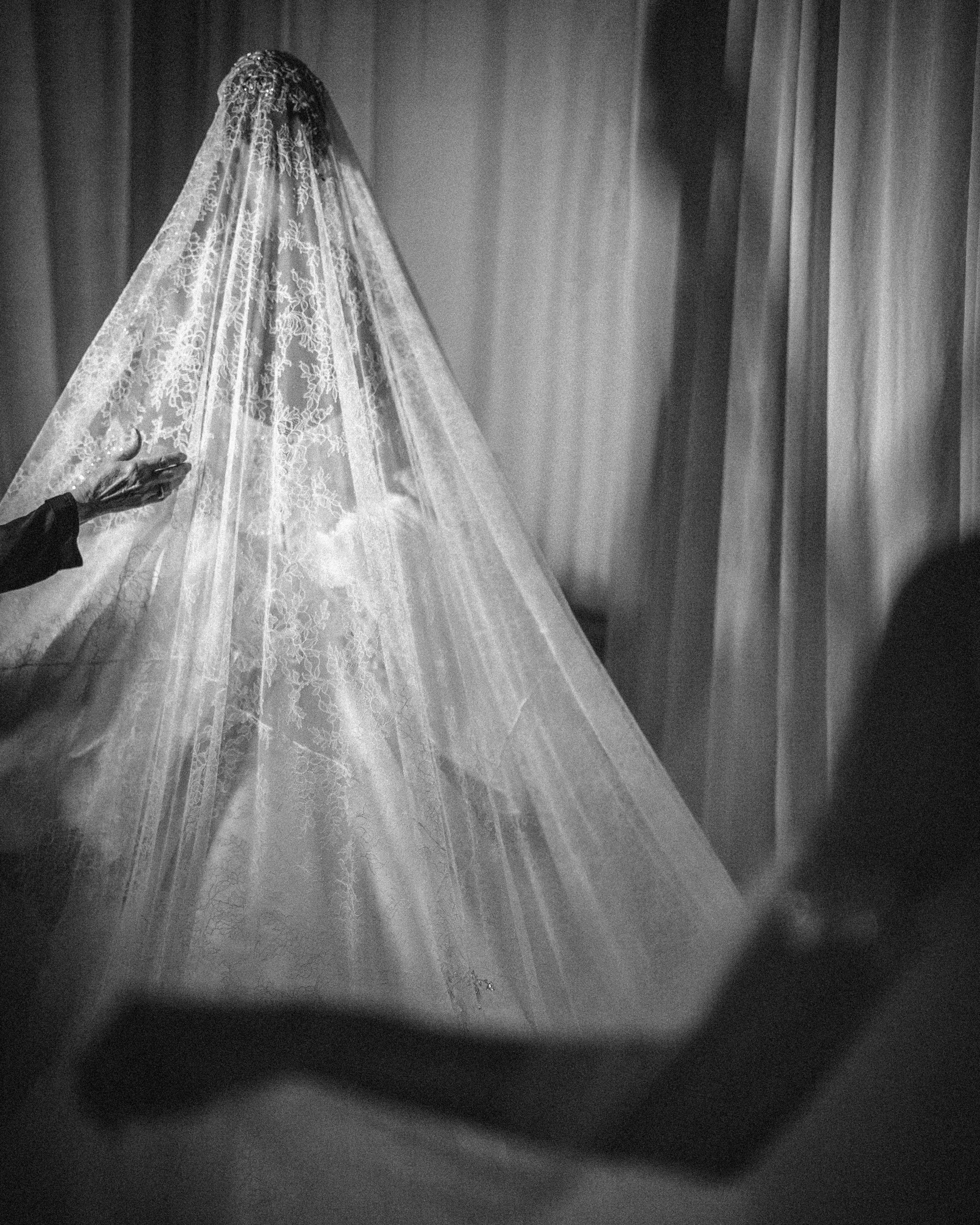 In a captivating black and white image, a person stands enveloped in a lace-covered garment, possibly from the Pnina Tornai AURA Collection. Bathed in soft lighting, they extend an inviting hand toward the viewer.