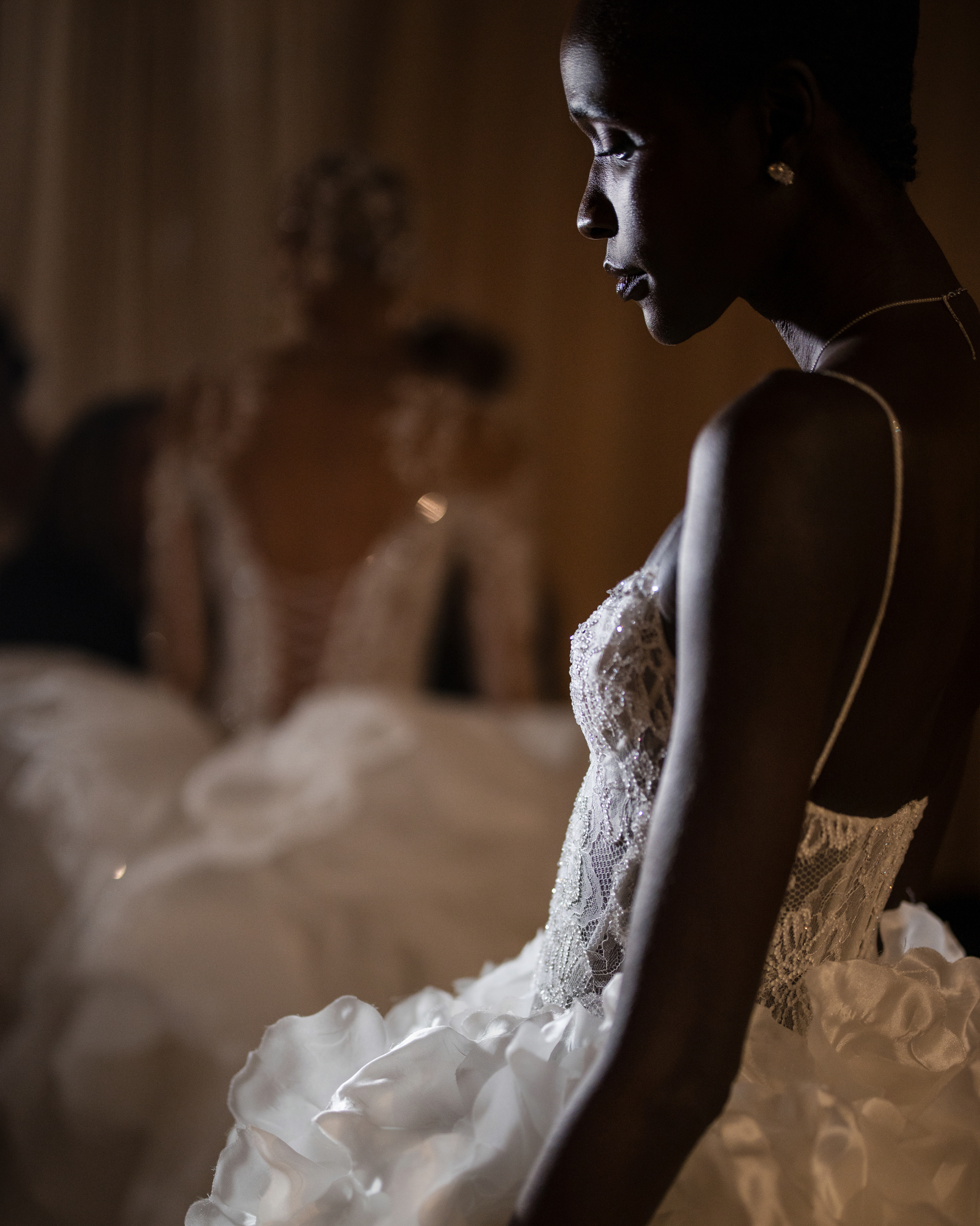 A model in a detailed white dress with a textured bodice from the Pnina Tornai AURA Collection stands in profile. Another model in a similar dress is softly out of focus in the background.
