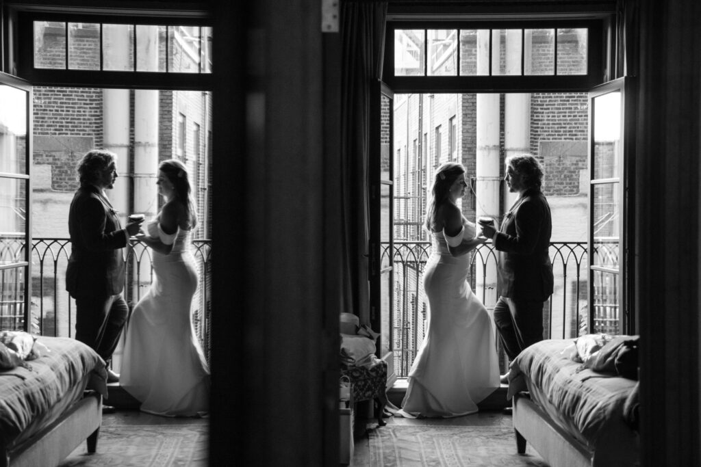 A couple stands in front of a window at the Greenwich Hotel wedding, holding drinks, their reflection visible in the glass. They're dressed in formal attire, the elegant setting exuding a serene indoor ambiance.