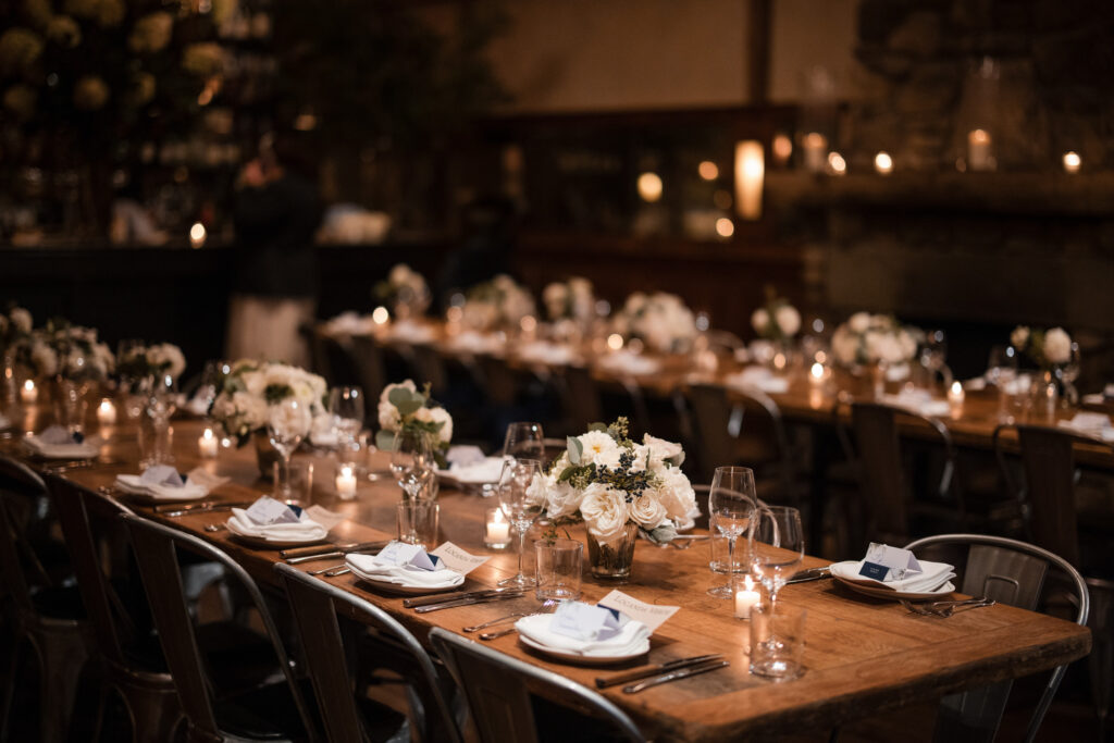 A dimly lit dining setup at Locanda Verde Wedding features elegantly arranged tables with white floral centerpieces, candles casting a gentle glow, and neatly arranged plates and glasses.