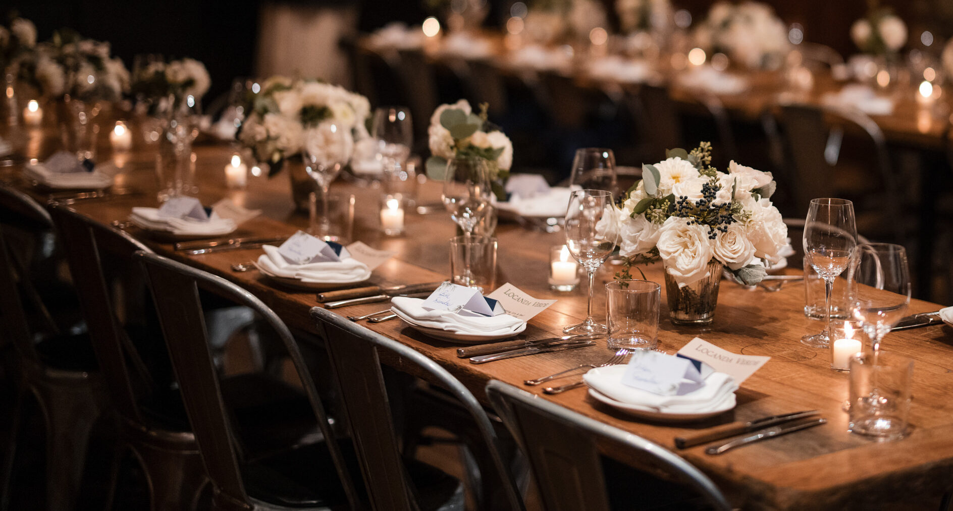 A dimly lit dining setup at Locanda Verde Wedding features elegantly arranged tables with white floral centerpieces, candles casting a gentle glow, and neatly arranged plates and glasses.
