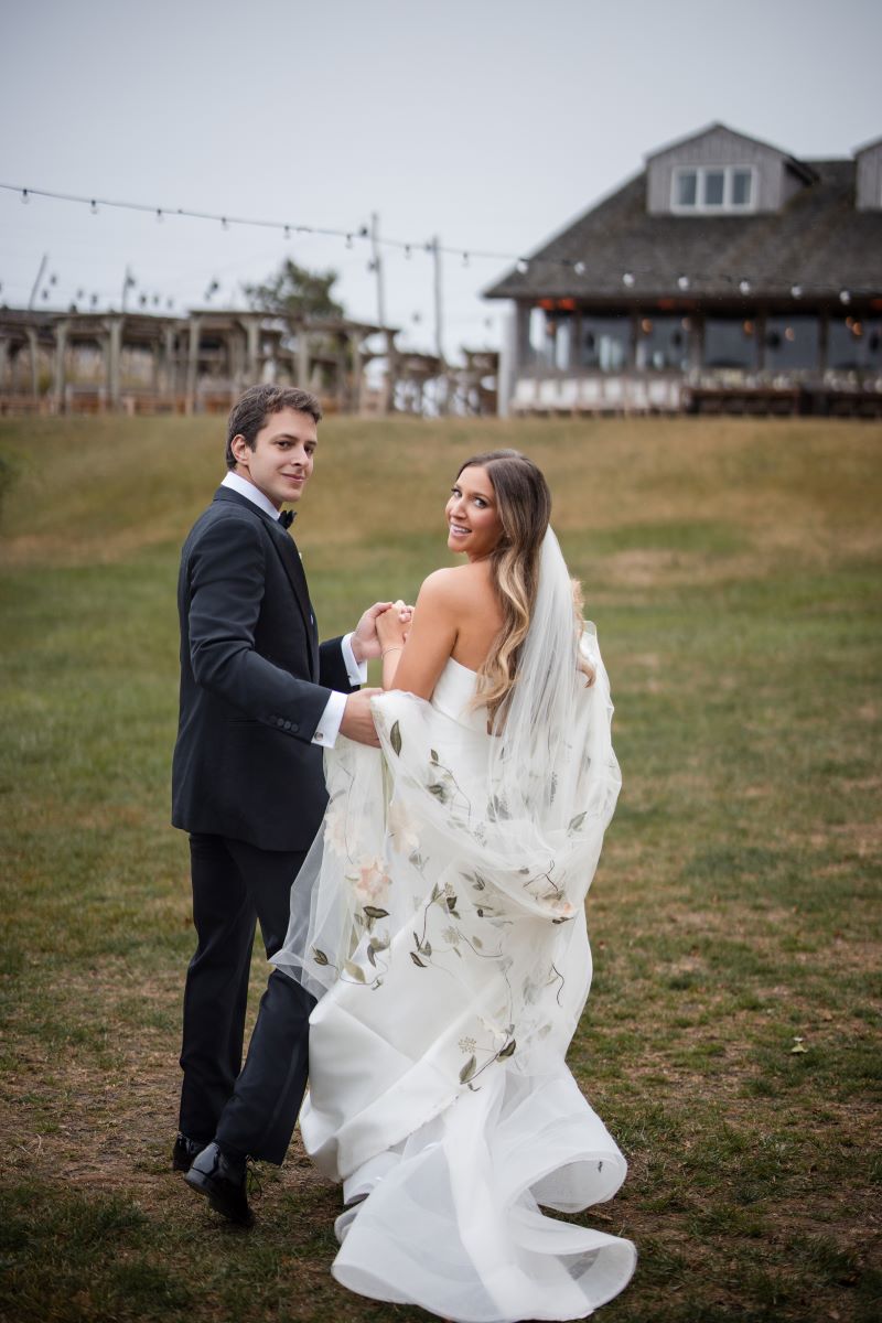 woman in a white wedding dress and a man in a suit standing in a yard and both are looking over their shoulders they are holding hands and the man is helping the woman with her wedding dress