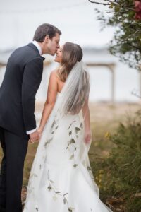 A woman in a white wedding dress and a man in a suit holding hands and the man is leaning in to give the woman a kiss