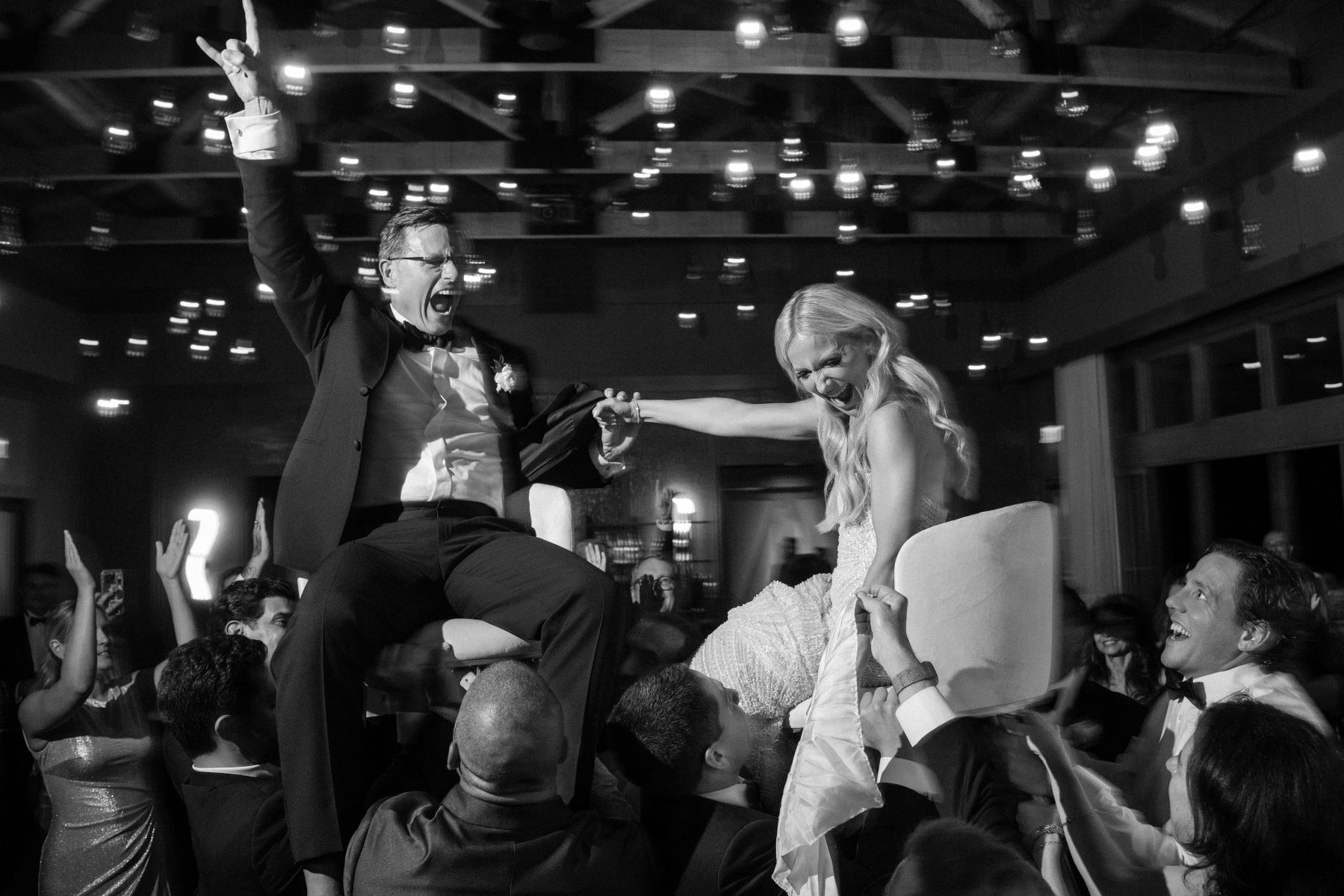 A married couple sitting in two chairs the charis are being lifted up into the air by their guests the woman is wearing a wedding dress and laughing and the man is wearing a tuxedo and pumping his fist and cheering with his guests the woman and the man are both holding hands 