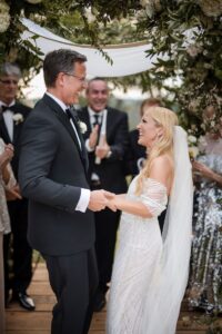 A woman in a white wedding dress and wedding veil is looking at her partner and laughing and holding his hands the man is in a suit and also is laughing as their guests clap and cheer standing behind them