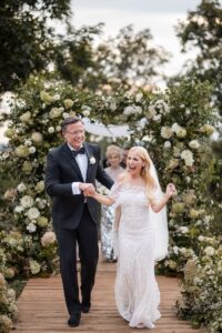 A woman is a white wedding dress is smiling and laughing and holding the hand of her partner who is wearing a suit and touching her back behind them is a floral arch