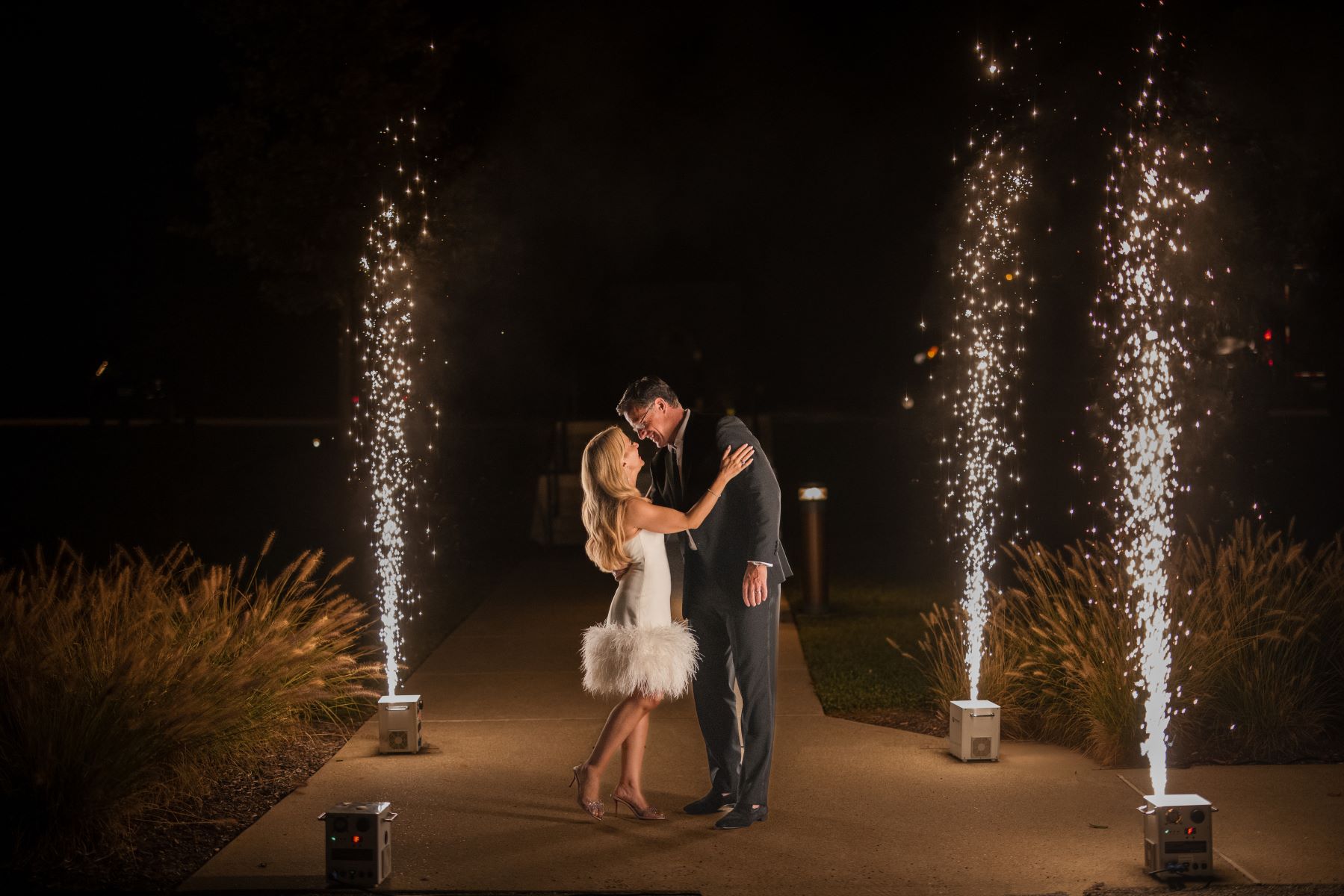 A woman in a short white dress and heels is touching her partner's shoulder and laughing the man is leaning towards her and is wearing a suit around them are wedding sparkler boxes