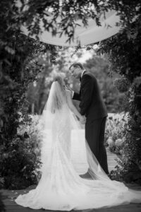 A man in a suit is gently touching the face of his partner who is in a white wedding dress and veil and both are looking at each other