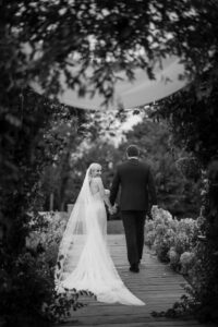 A woman in a wedding dress with a veil is looking back over her shoulder and holding the hand of her partner who is wearing a suit and is walking fowards they are surrounded by tress and bushes