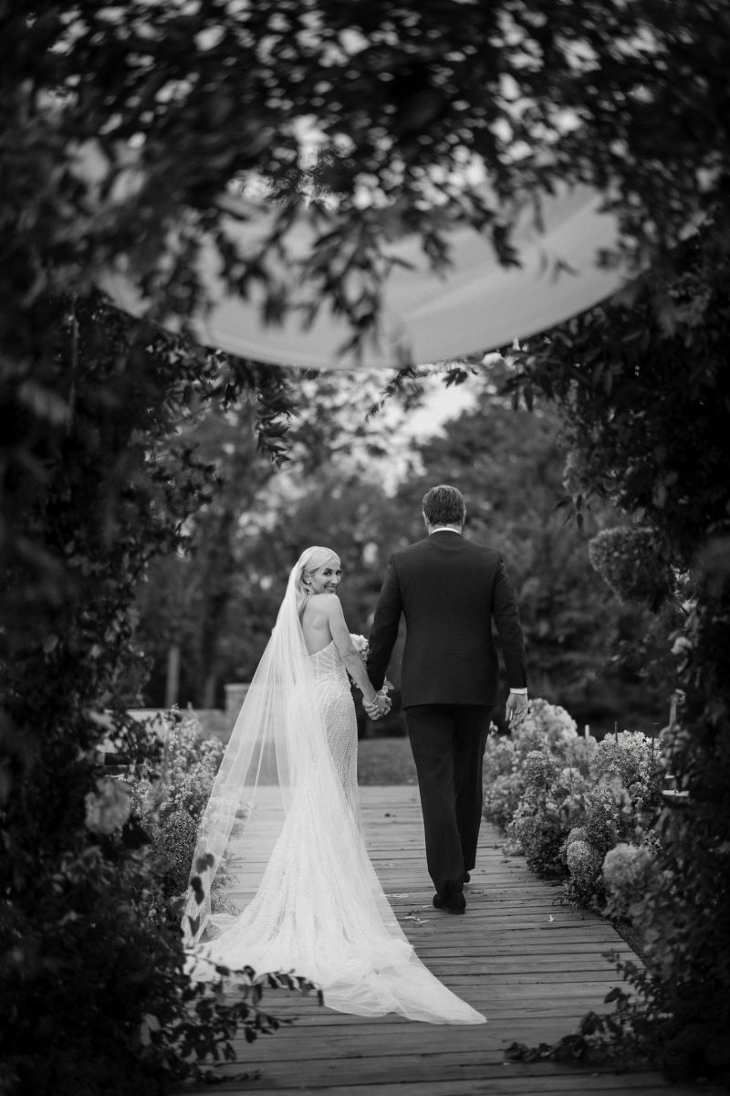 A woman in a wedding dress with a veil is looking back over her shoulder and holding the hand of her partner who is wearing a suit and is walking fowards they are surrounded by tress and bushes