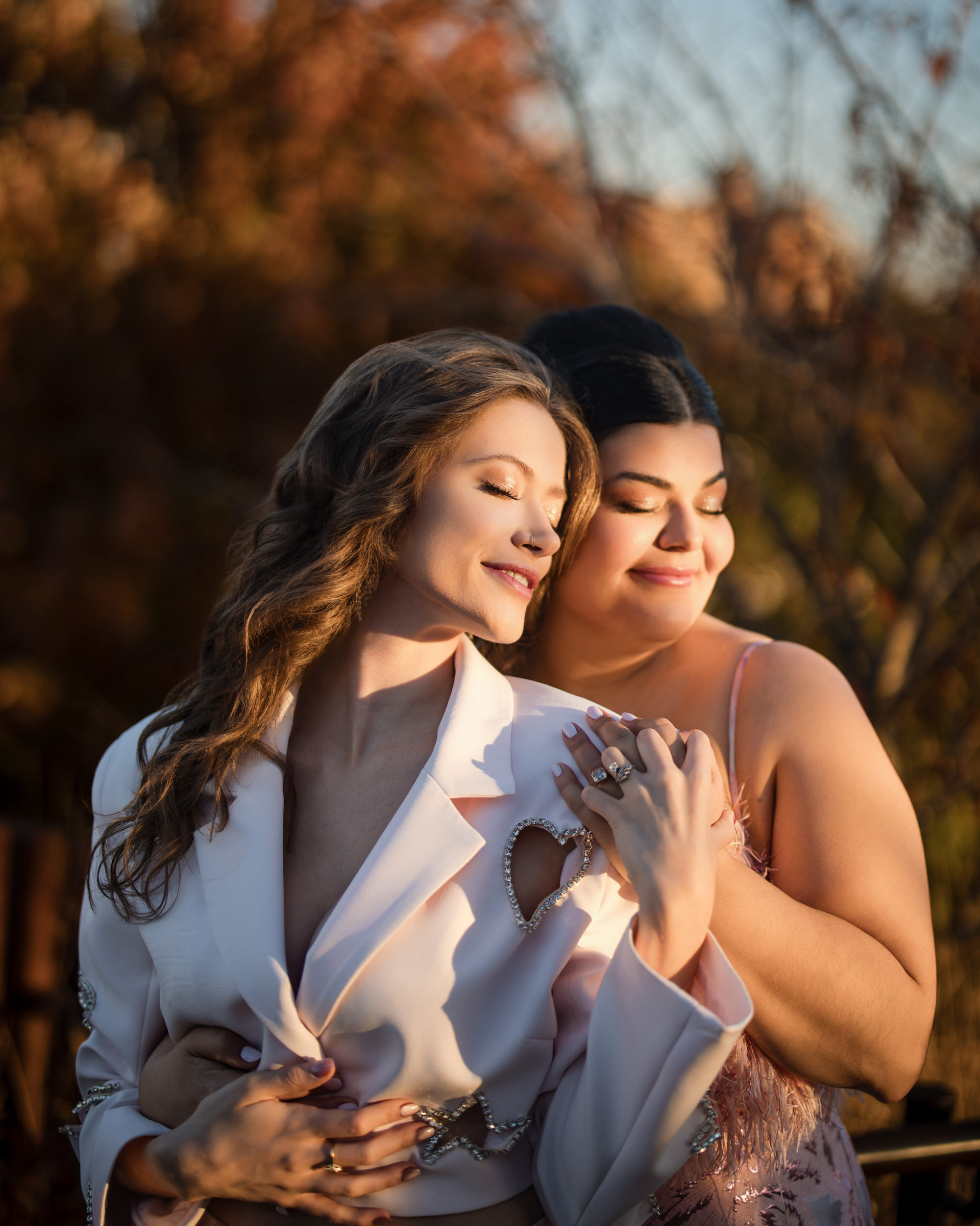 Two women, one wearing a white jacket and the other in a pink dress, embrace outdoors on Little Island with eyes closed, surrounded by warm, golden light at their engagement photo session.