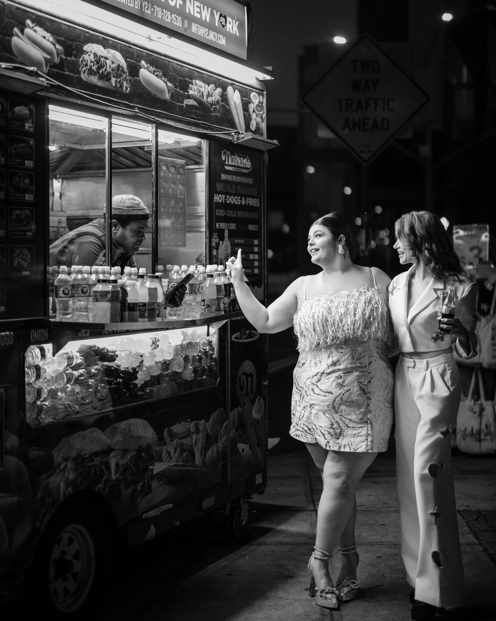 Two women in elegant evening attire stand by an NYC hot dog cart at night, interacting with the vendor. The scene, captured as an engagement photo, is beautifully rendered in black and white, adding a timeless charm to the fleeting moment on the bustling street.