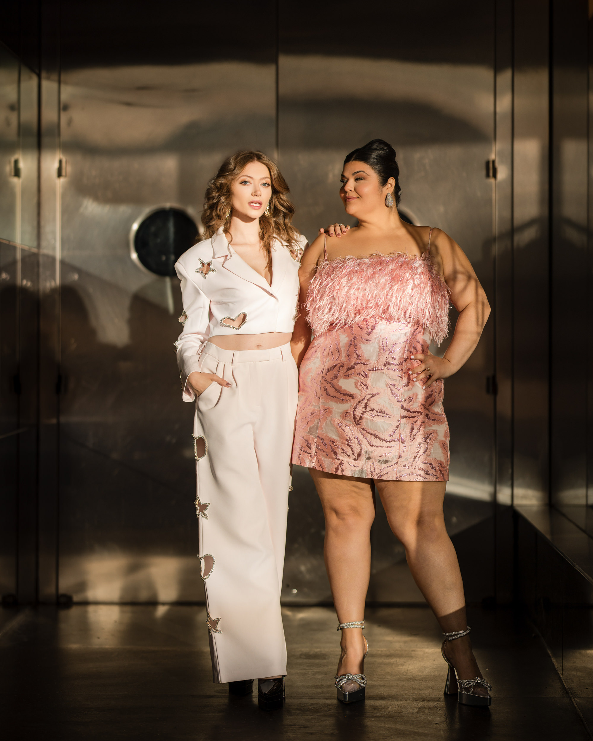 Two women pose stylishly; one in a white outfit with star cutouts, the other in a pink fringe dress, capturing the edgy vibe of an engagement photo shoot. They stand against a metallic background that channels the essence of the Meatpacking District.