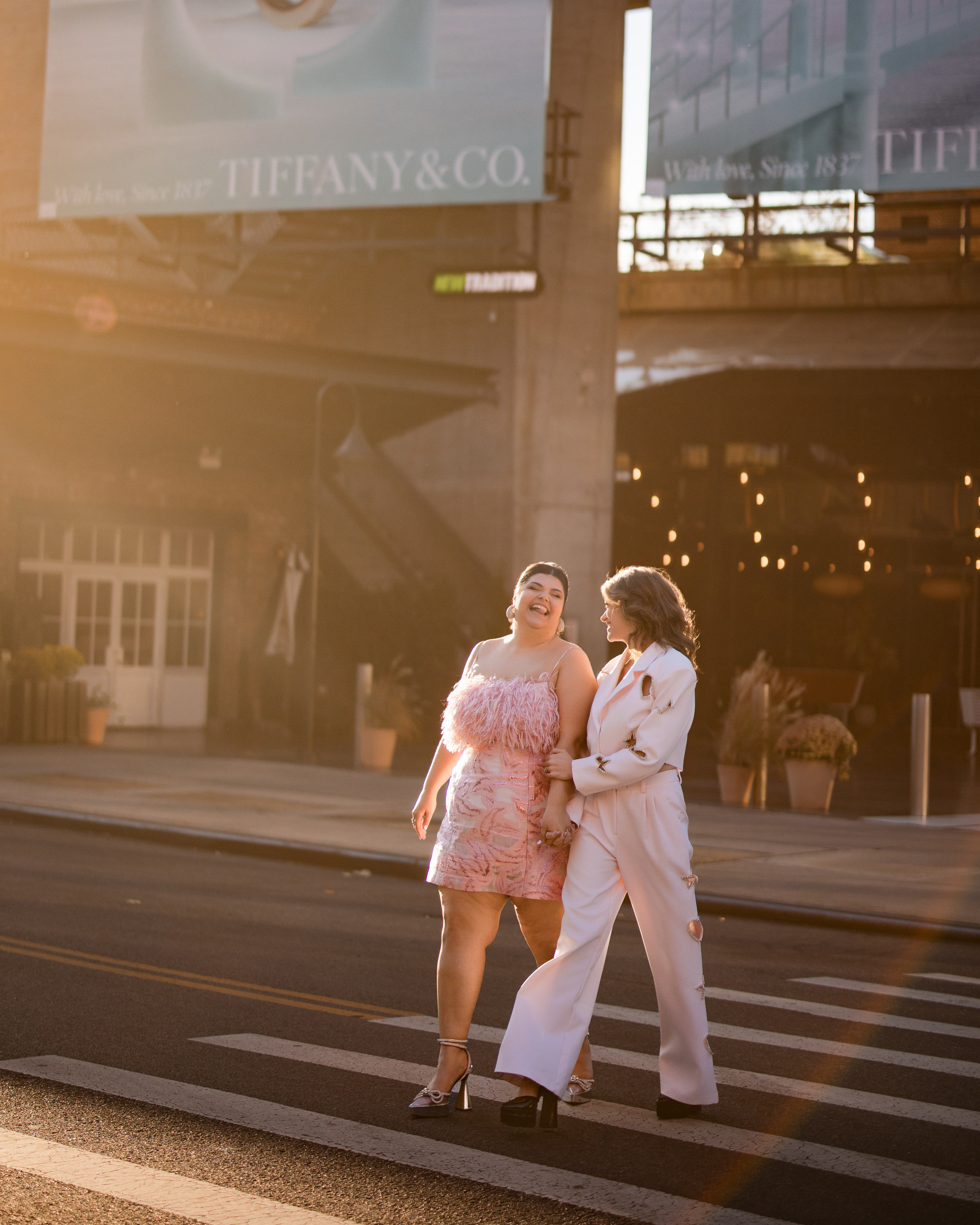 Two brides walking arm in arm across a city street, wearing stylish outfits—one in a pink dress with feather details, the other in a white suit. Sunlight streams in, creating a warm ambiance reminiscent of the standard Meatpacking engagement session.