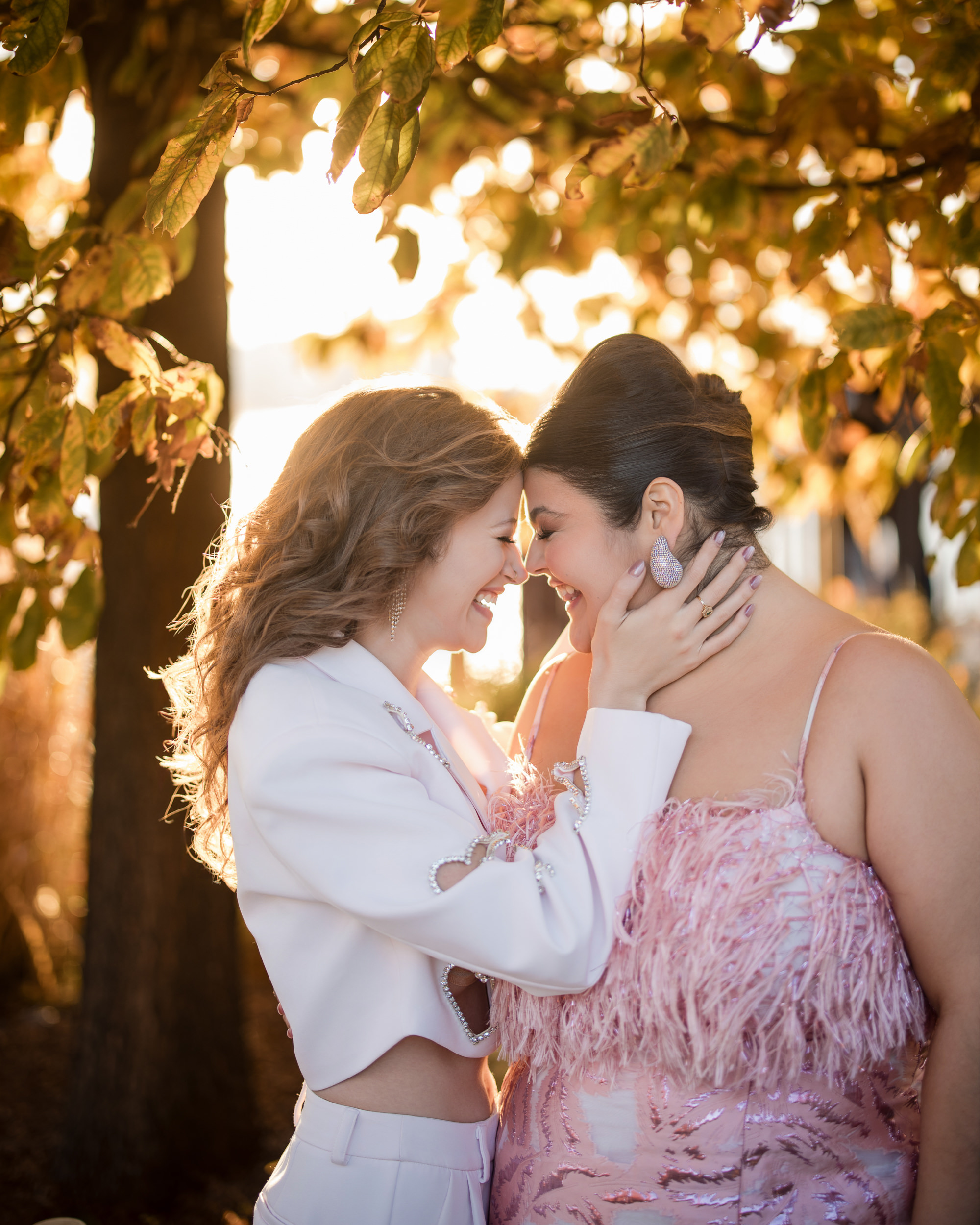 Two women, bathed in the warm hues of autumn leaves, smile and touch foreheads under a tree. One wears a white outfit, the other a pink dress—a tender moment that feels like an intimate engagement on their own little island of love at their engagement photo session.