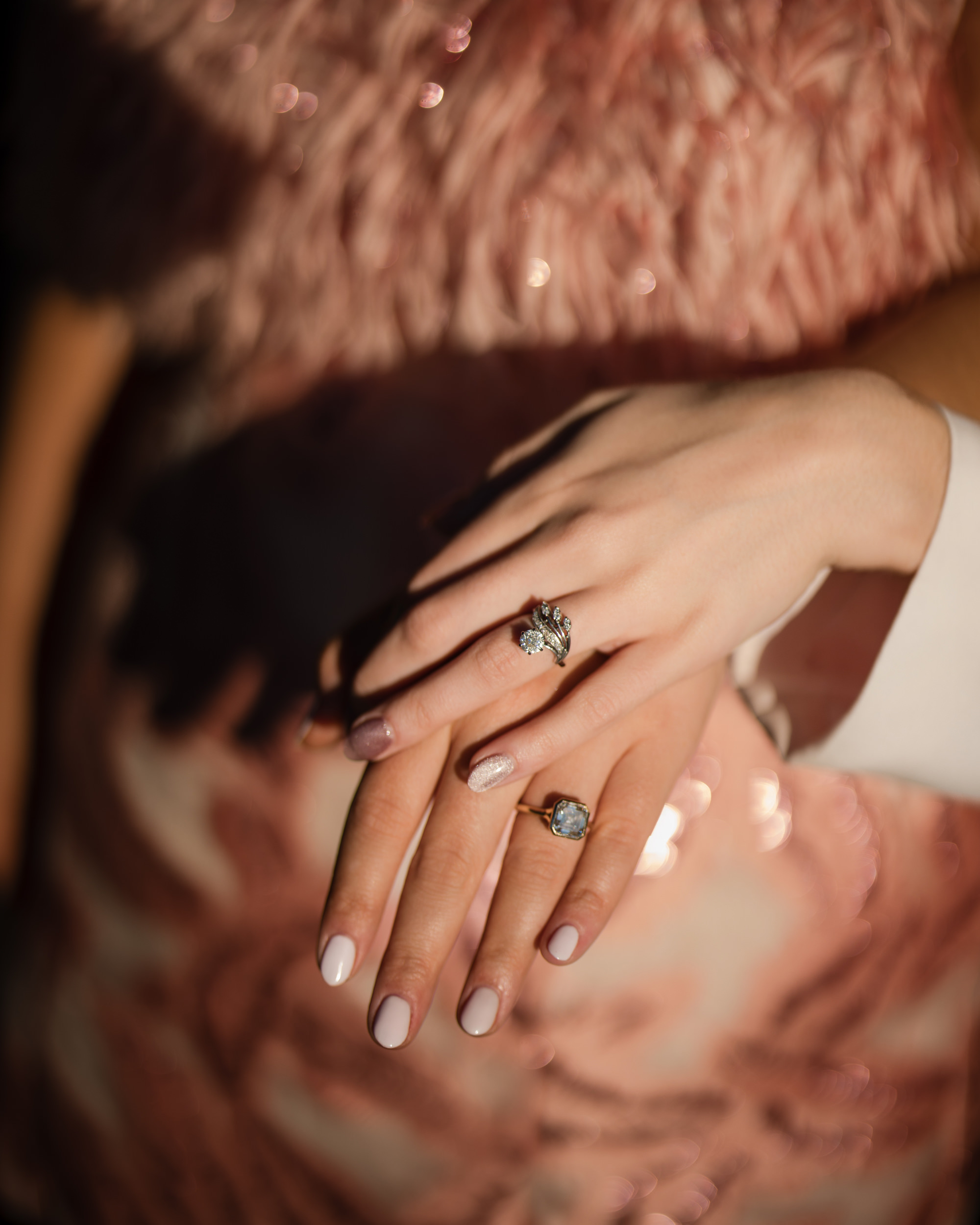 Close-up of two hands with manicured nails, wearing stunning diamond rings that sparkle like the stars over a little island sky, placed one over the other against a sequined pink fabric background.