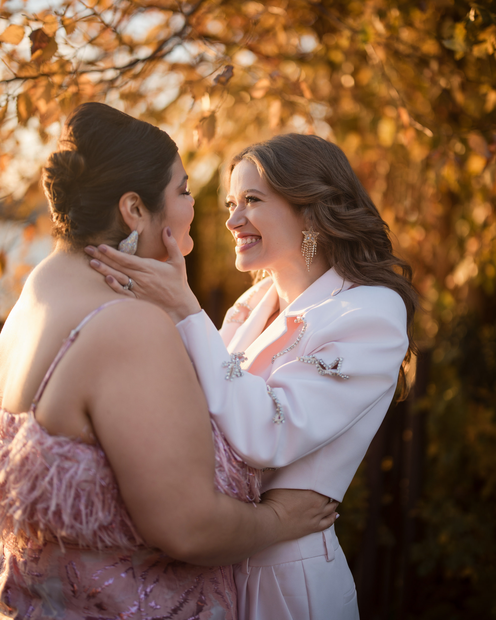 Amidst a tapestry of autumn leaves, two people share smiles—one in a crisp white suit, the other in a flowing pink dress. Their joy mirrors that of little island engagement memories cherished forever.