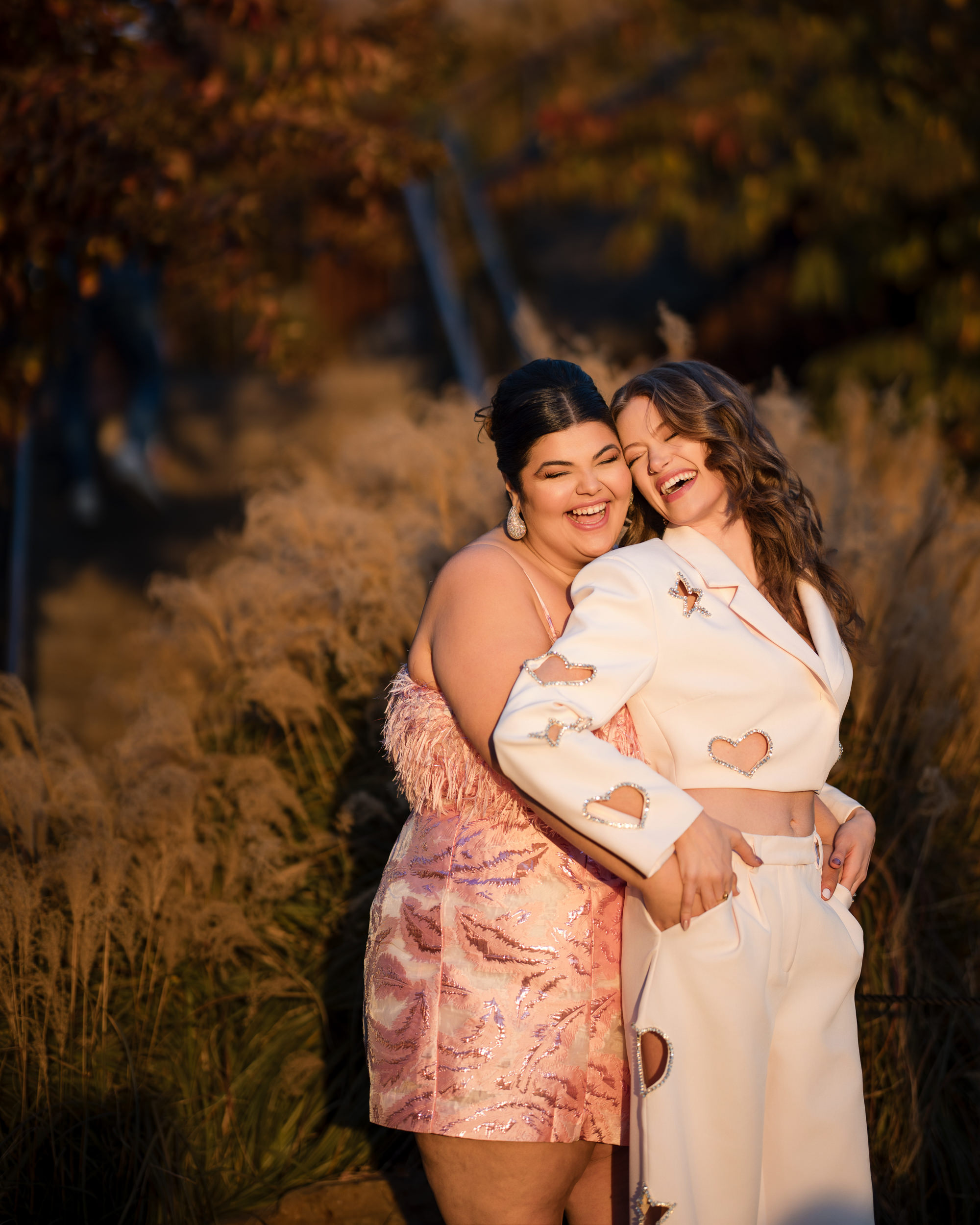 Two women smile and embrace in a sunlit outdoor setting, surrounded by tall grass and blurred foliage, celebrating their little island engagement.