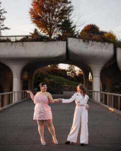 Beneath the bridge, two women stand hand in hand, celebrating their little island engagement. One is dressed in a pink dress, while the other dazzles in white attire. Behind them, the trees sway gently, adding a touch of nature's charm to their special moment.