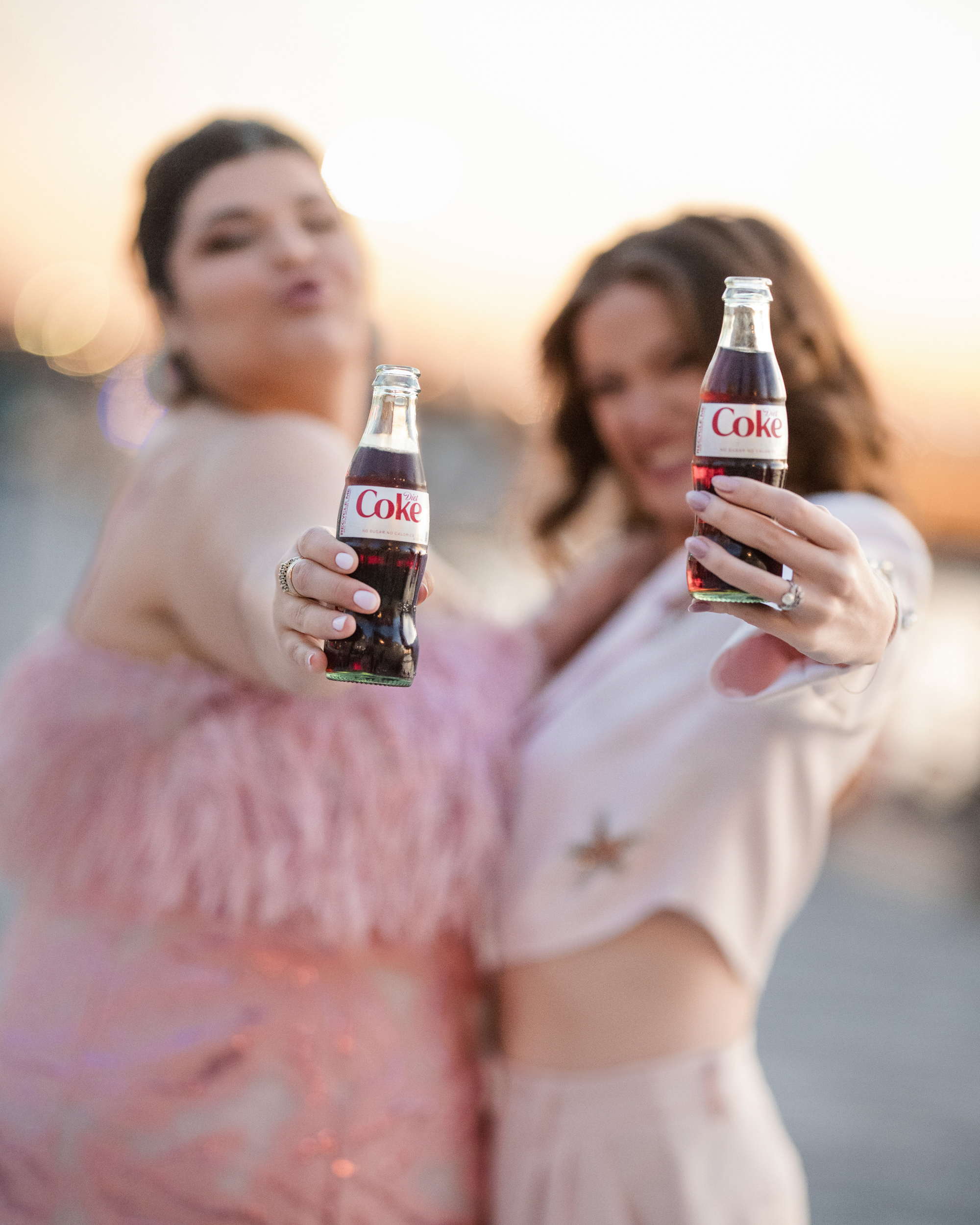 Two women in festive attire hold Diet Coke bottles toward the camera, smiling at sunset on a little island, celebrating their engagement.