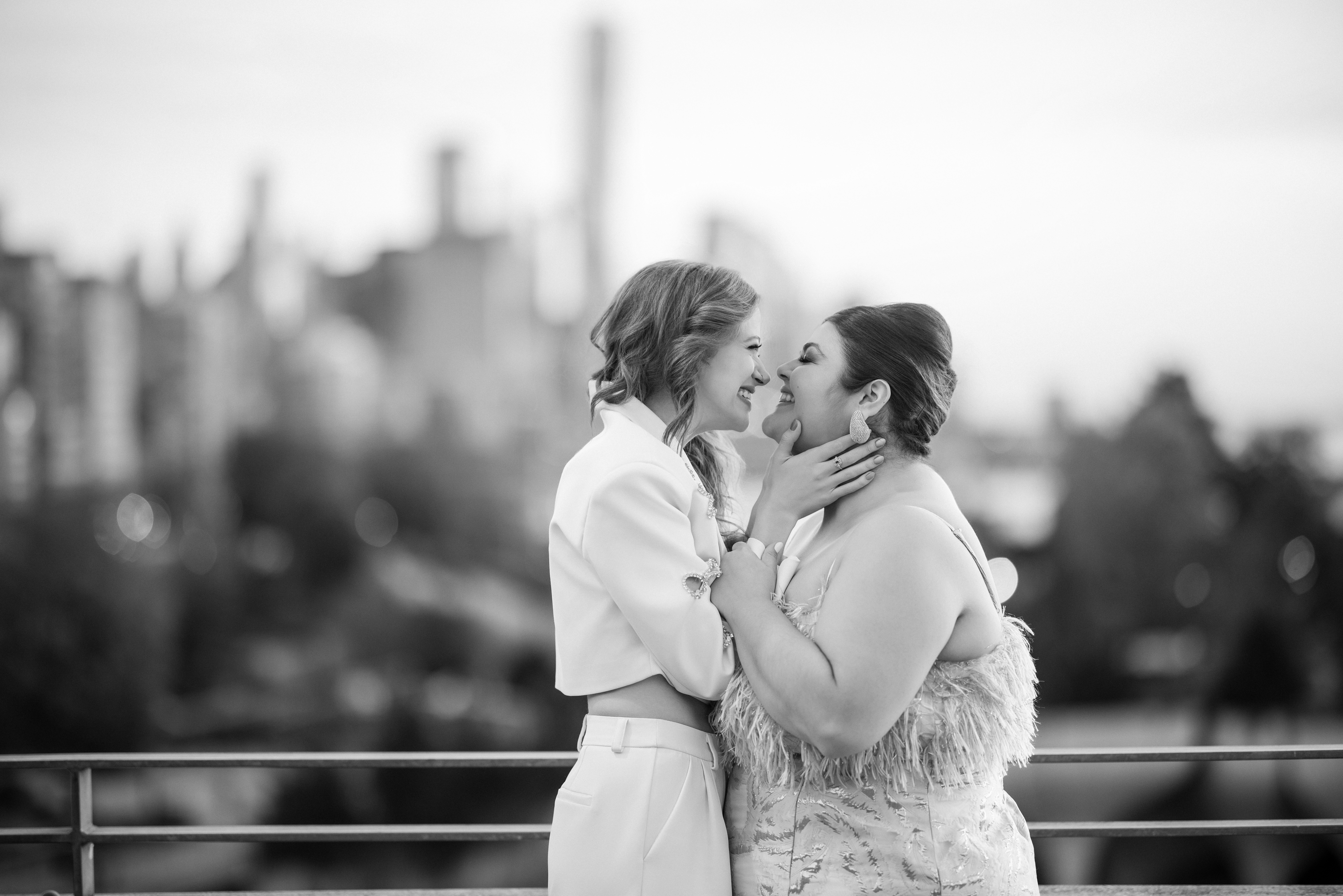 Two women, celebrating their engagement, smile at each other on a rooftop, with the cityscape and hints of Little Island NYC blurred in the background.