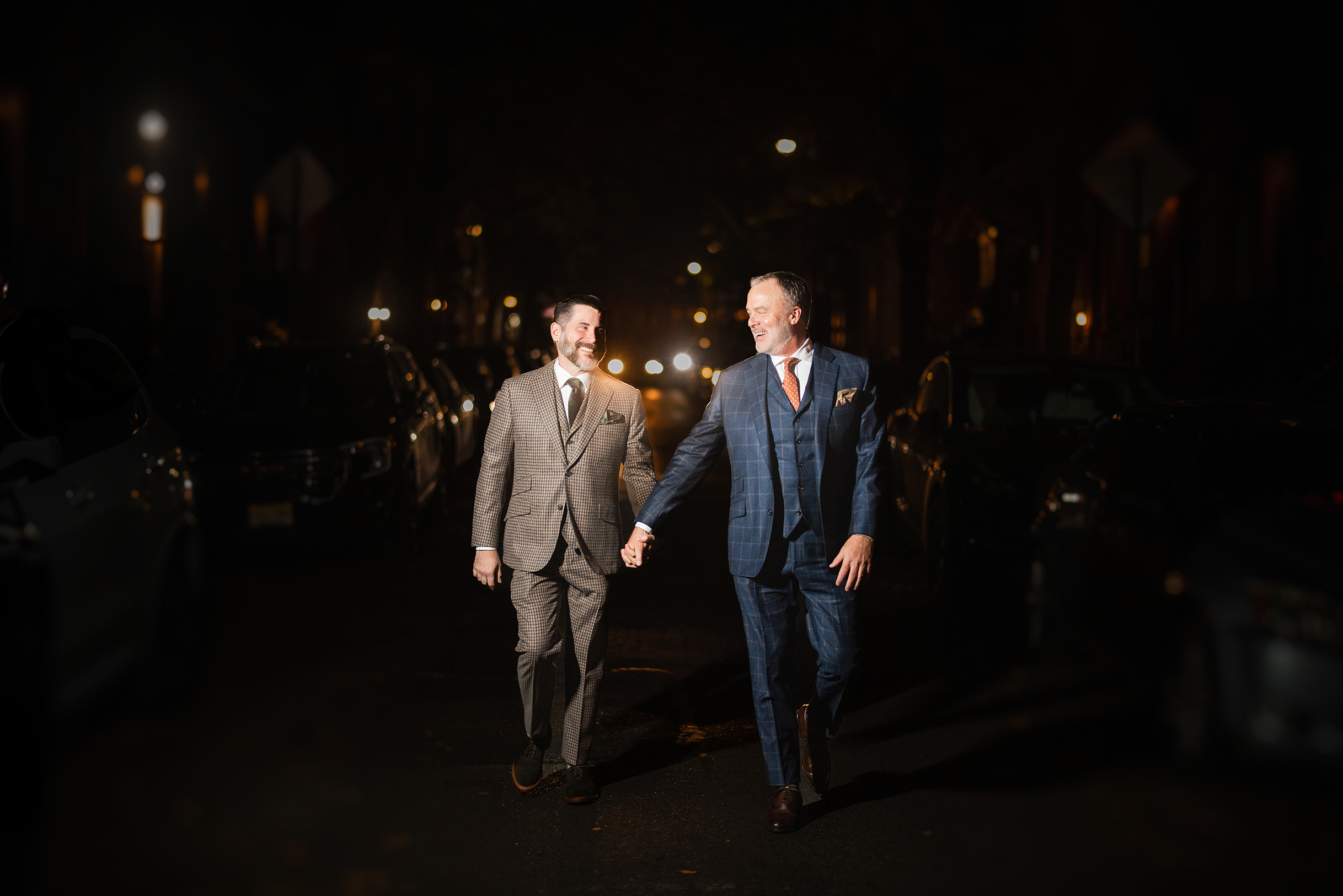 Two men in suits holding hands, walking on a dimly lit street at night near The Townhouse, smiling at each other with the warmth of a little owl's embrace.