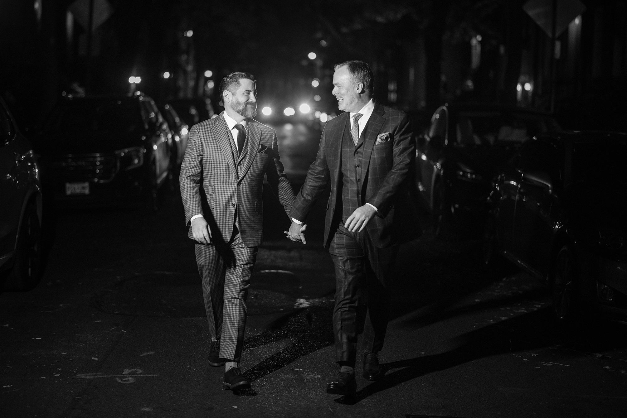 Two men in suits walk hand-in-hand on a street at night, illuminated by headlights from The Townhouse wedding venue in the background.