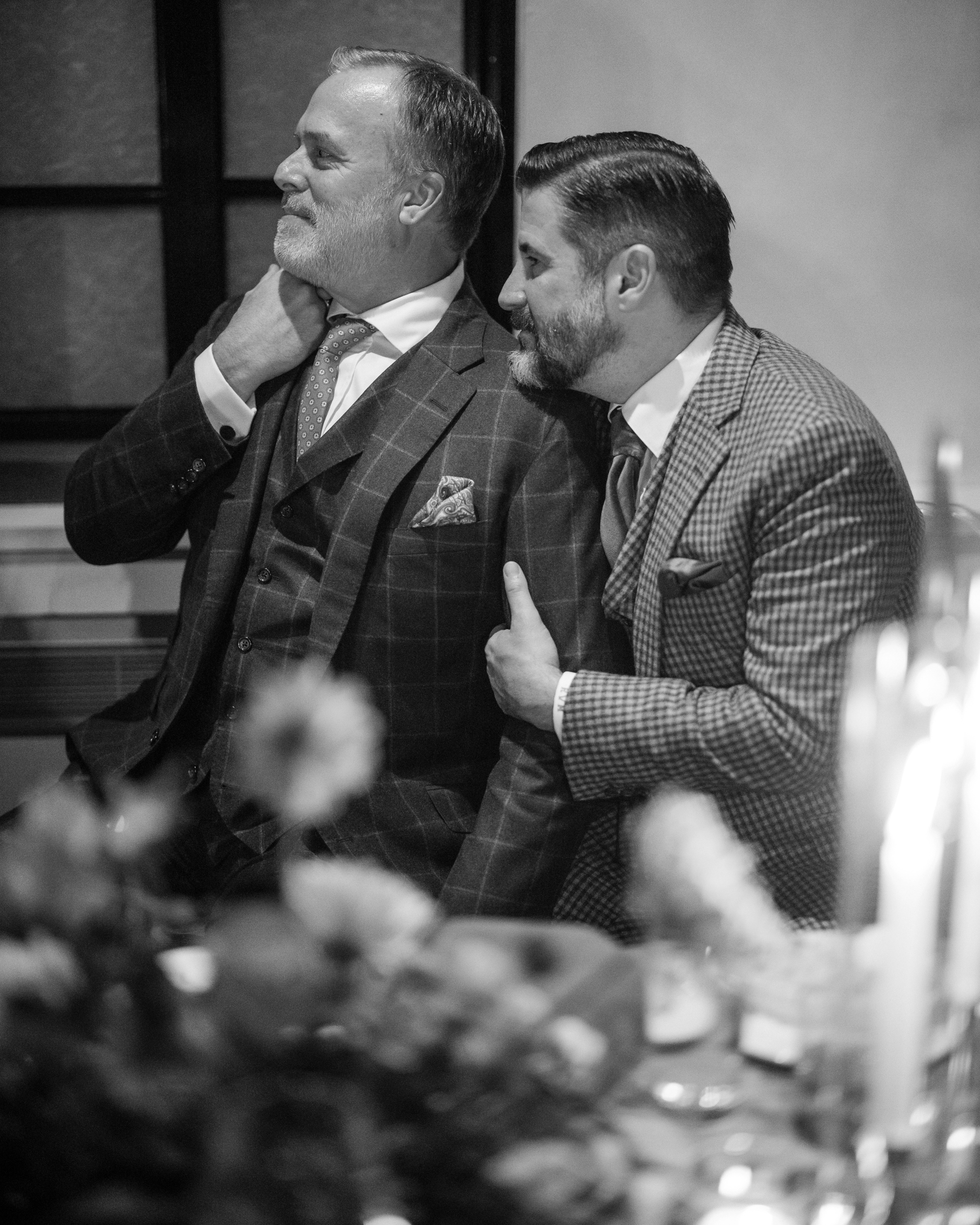 Two men in suits sit closely together at a table, one adjusting his tie and the other placing a hand on his shoulder. The intimate black-and-white scene exudes the charm of a townhouse wedding, capturing a moment reminiscent of wisdom shared between old friends, like whispers from a little owl.