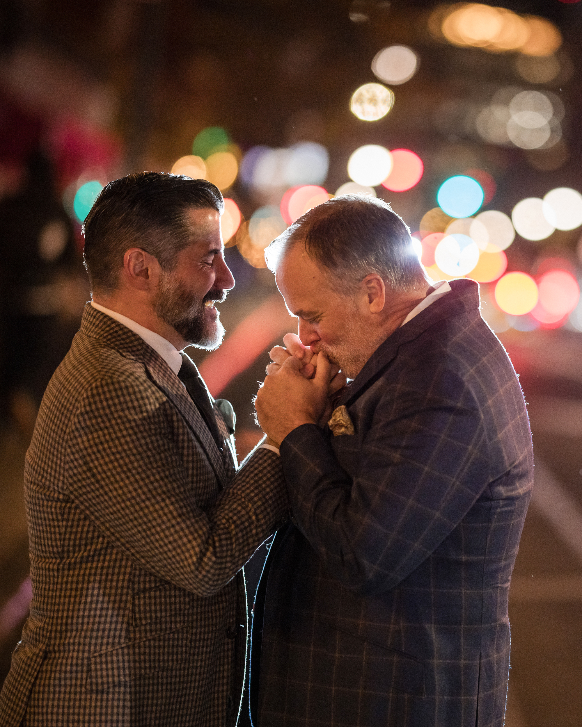 Two men in suits stand close on a city street at night, with one gently holding the other's hand outside The Townhouse. Colorful city lights blur in the background, reminiscent of a little owl's watchful gaze over their tender moment.