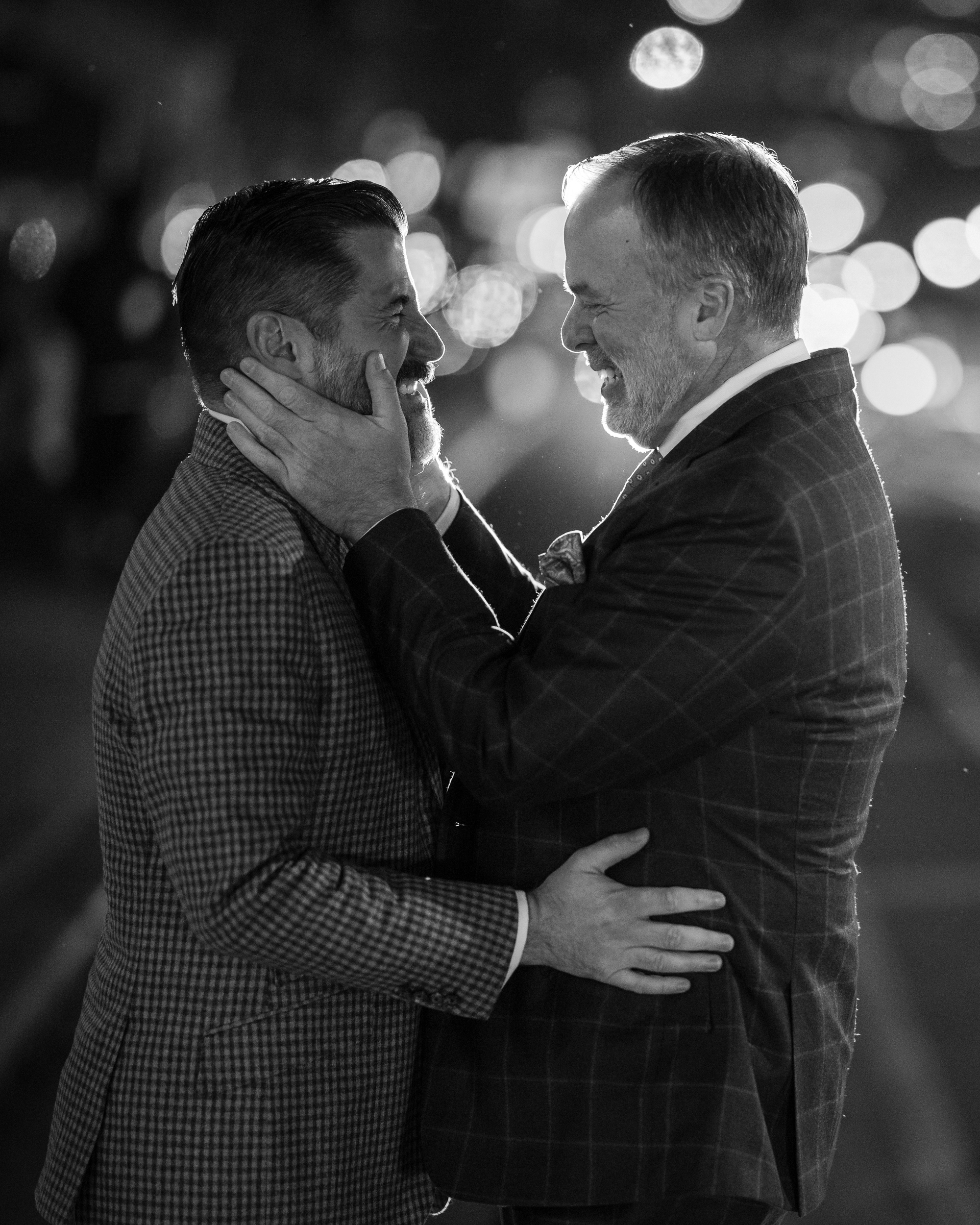 Two men in suits stand closely on a city street at night, gazing at each other affectionately like a scene from a Little Owl wedding, with blurred lights in the background.