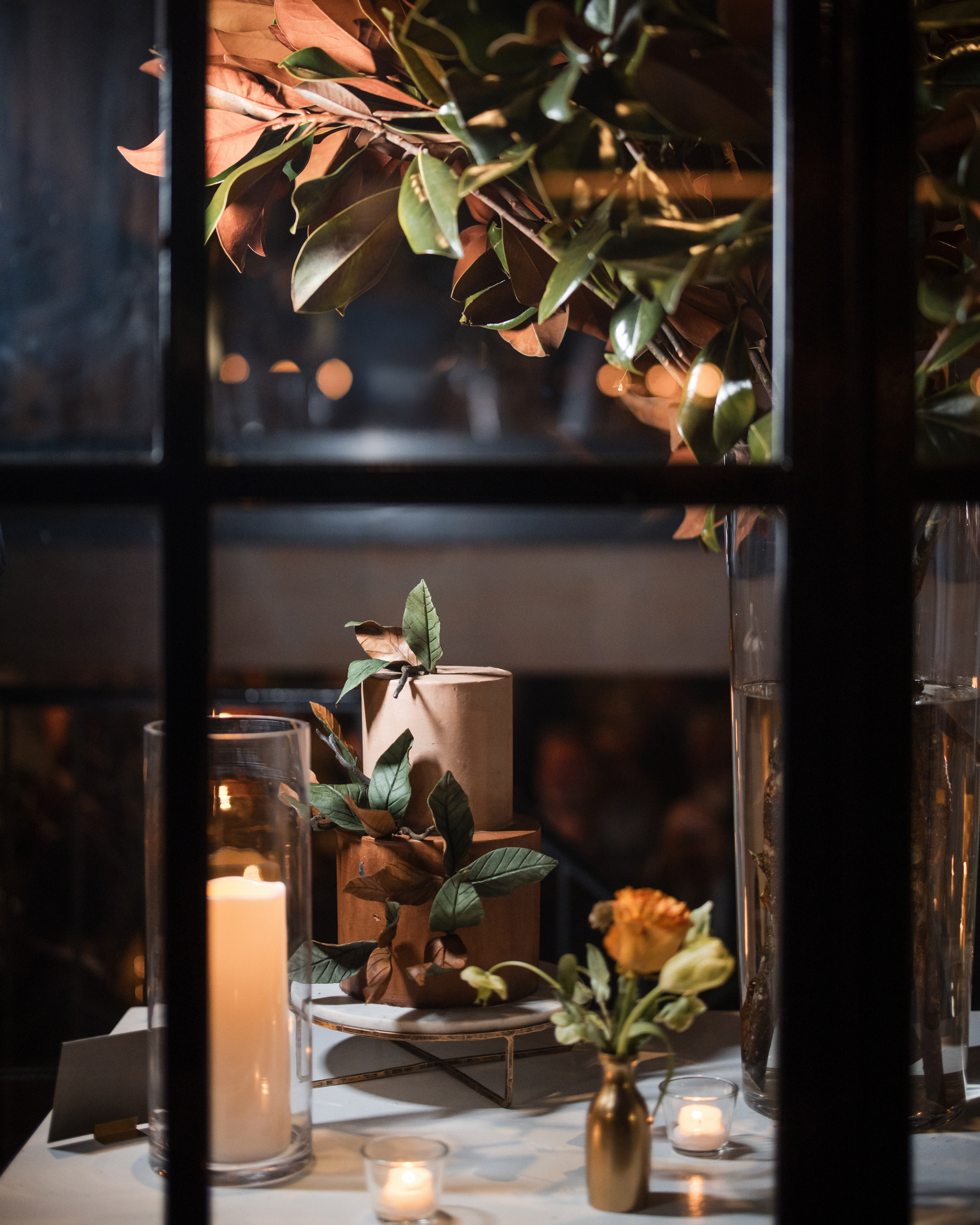 A cake adorned with foliage and a little owl decoration sits elegantly on a table surrounded by candles and a yellow rose in a vase, creating an enchanting scene glimpsed through the window panes at the townhouse wedding.