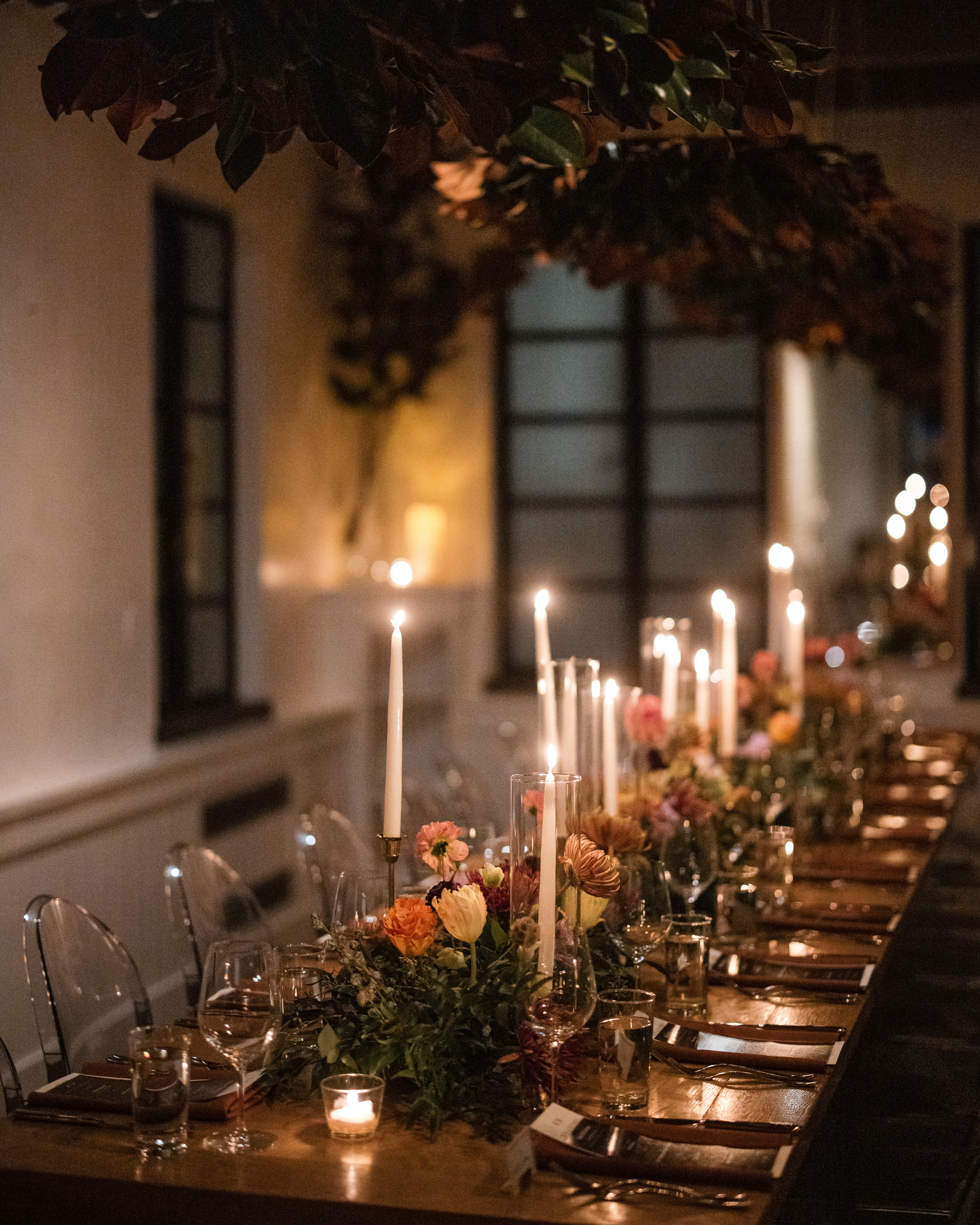 A dimly lit dining table at Little Owl The Townhouse, adorned with flickering candles, floral centerpieces, clear chairs, and elegant place settings, creates the perfect ambiance for a wedding or formal dinner.