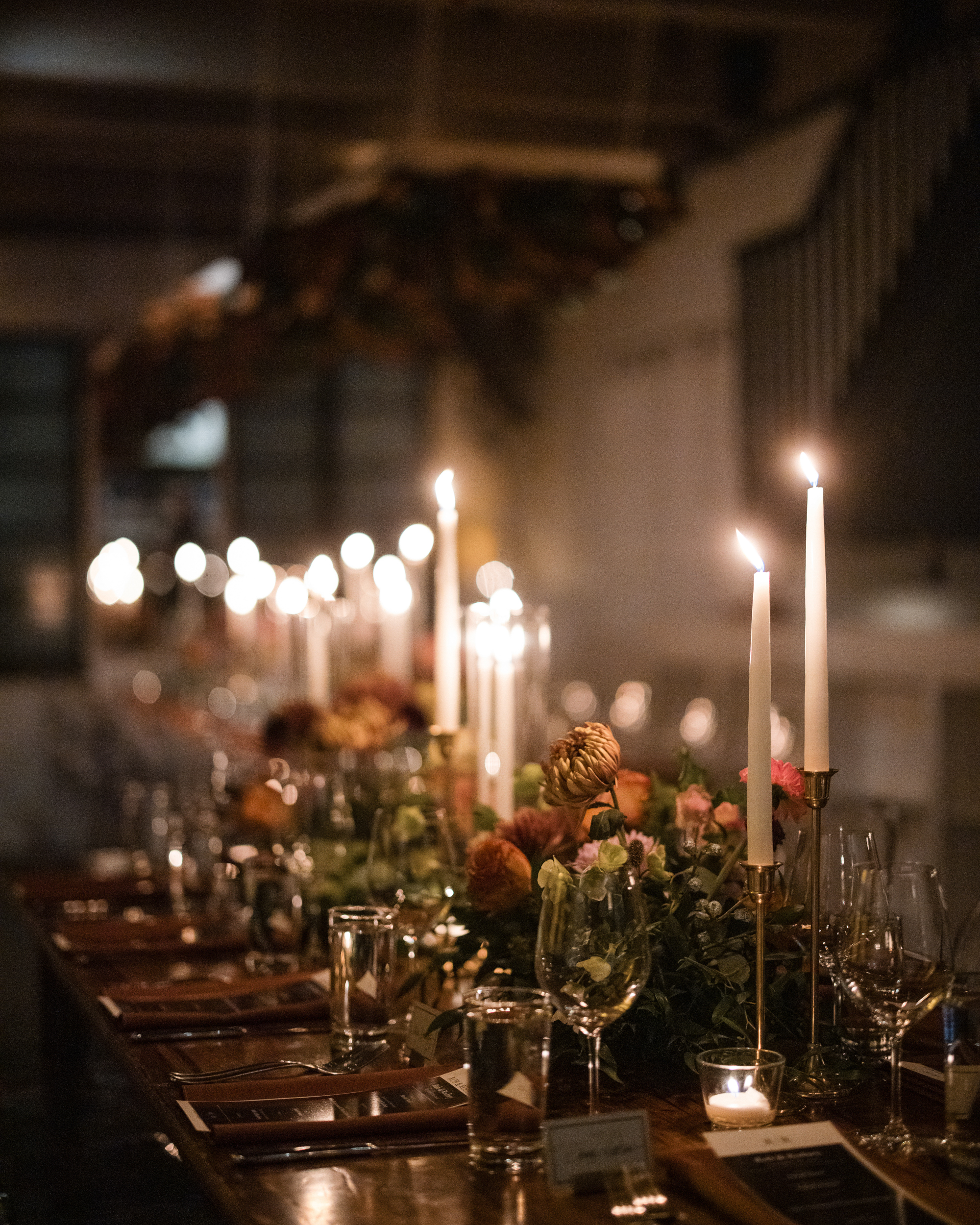 A long dining table is elegantly set with lit candles, glassware, and floral centerpieces at Little Owl The Townhouse, creating a warm and inviting atmosphere in the dimly lit room—a perfect setting for an intimate wedding celebration.
