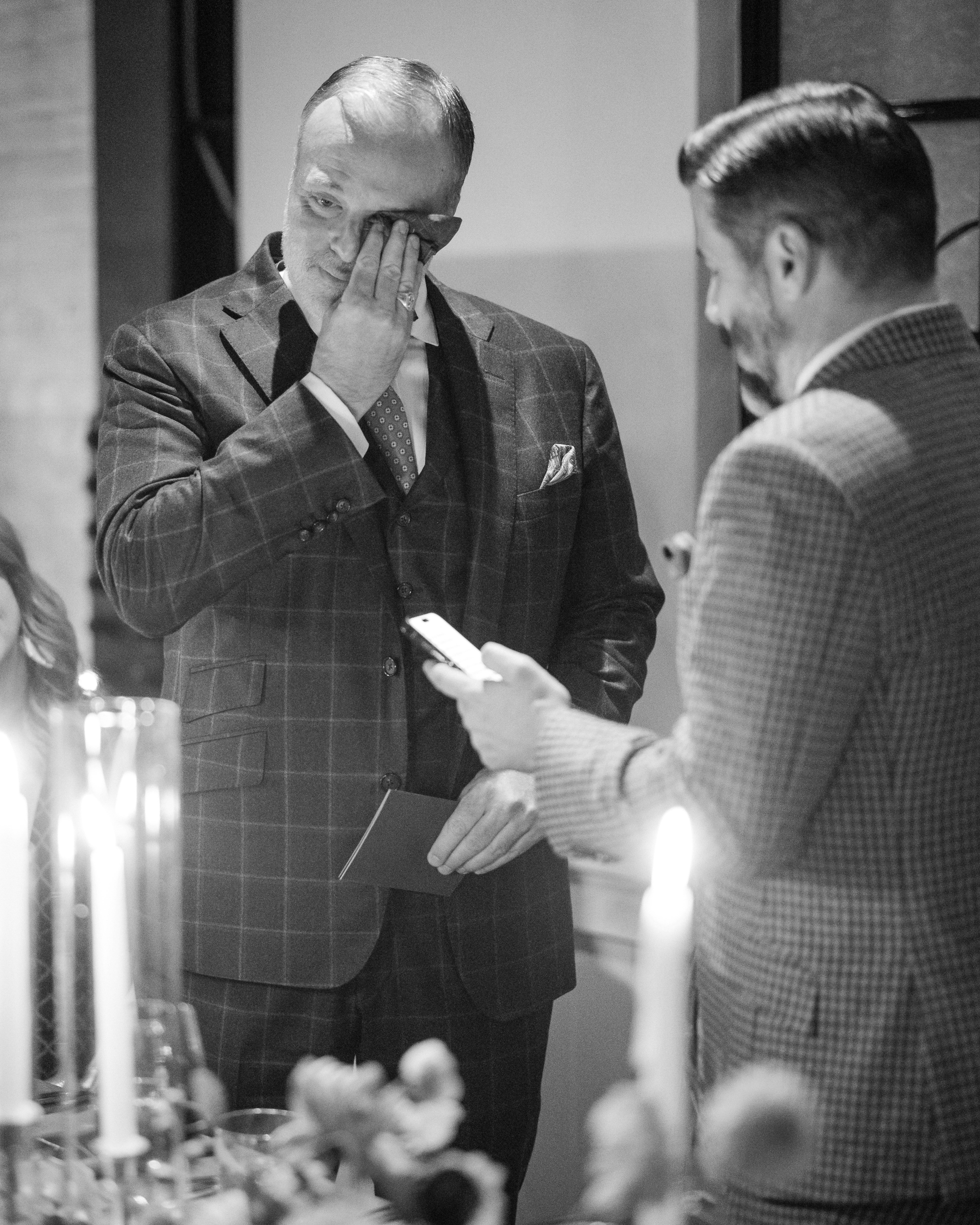 A man in a checkered suit wipes his eye as another talks on the phone, possibly sharing moments from a Little Owl wedding. Candles and blurred objects softly illuminate The Townhouse, adding to the heartfelt ambiance.