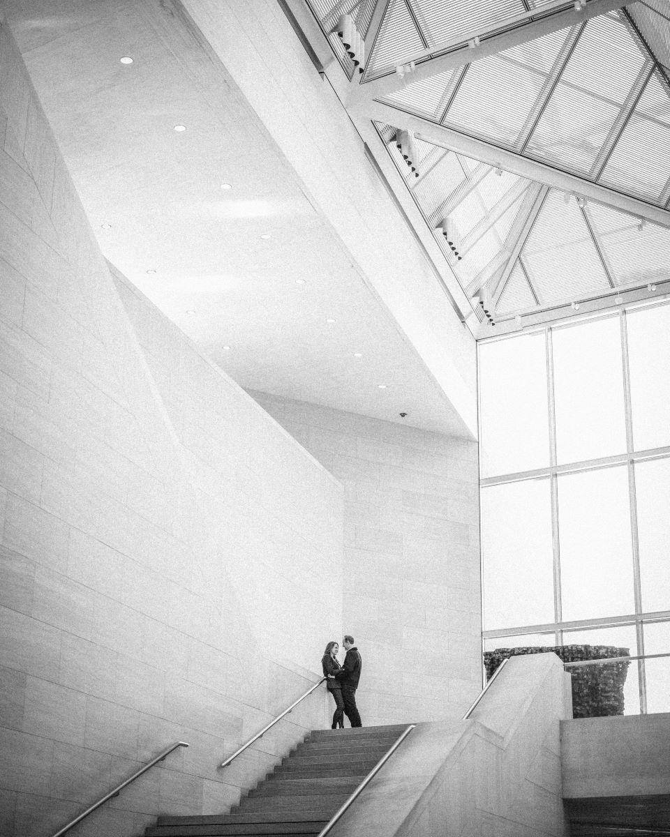 A couple at the top of a staircase next to large windows in a museum the woman is leaning against a rail and her partner has his hands on her hips 