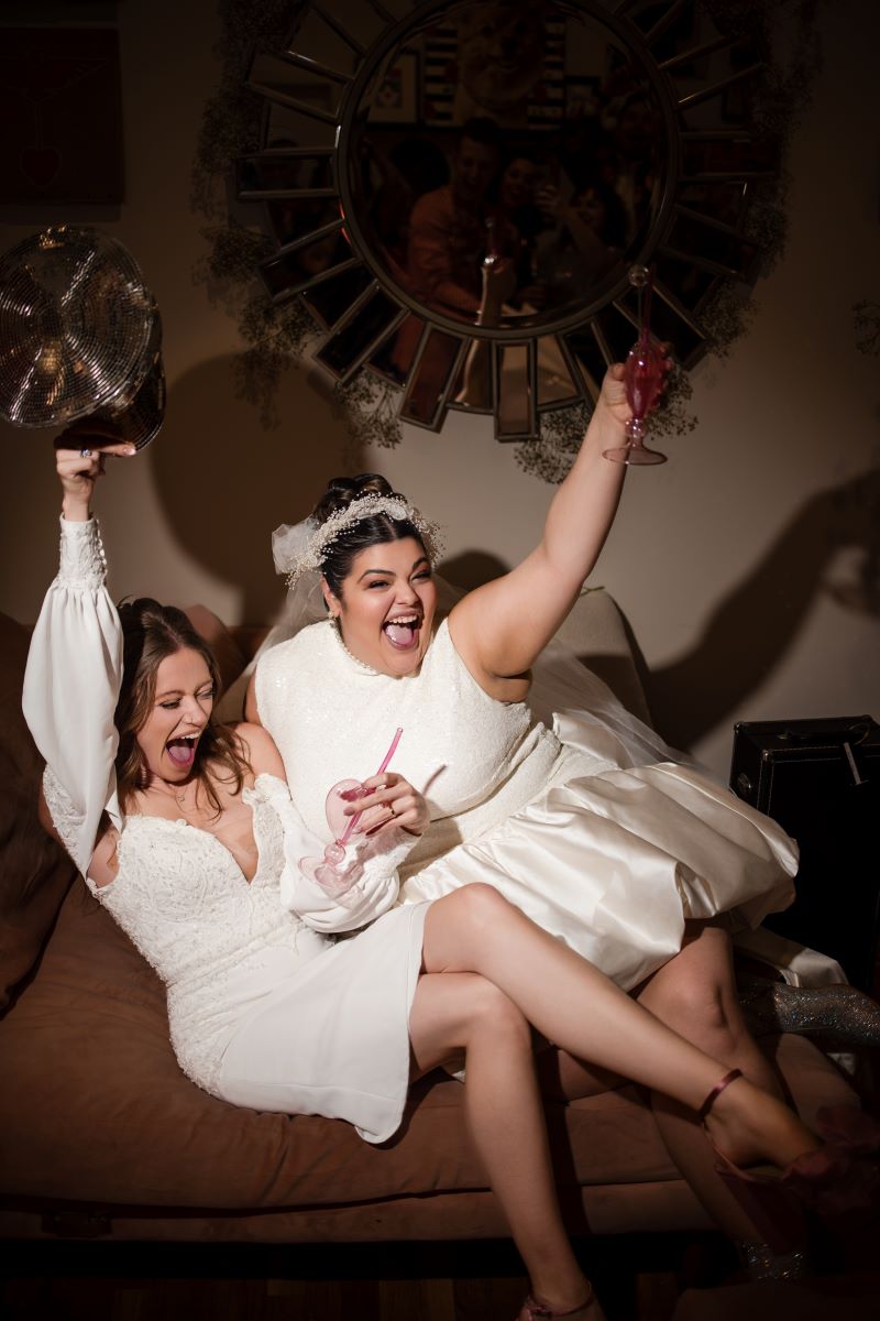 Two women in white dresses celebrating their wedding. They are both sitting on a couch, holding pink drinks and cheering