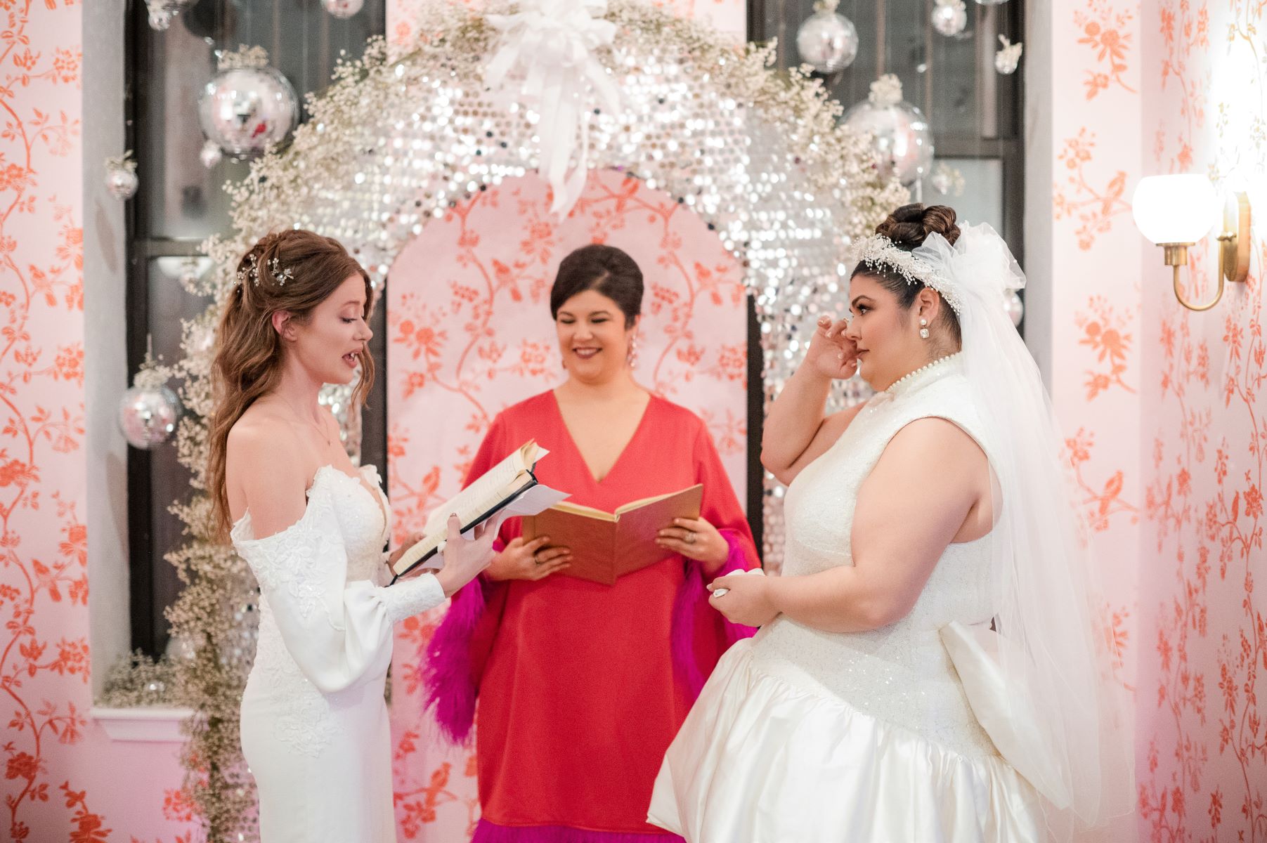 two women at their wedding ceremony in their home one woman is reading vows from a book and their officiant is standing behind them