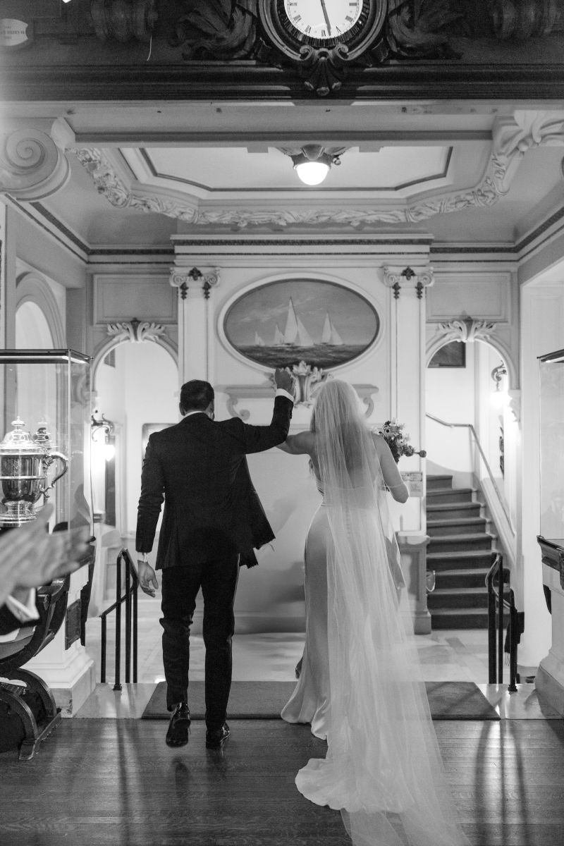 a woman in a white wedding dress and veil and a man ina tuxedo are about to walk down a staircase they are holding their hands in the air