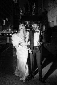 A couple in formal attire, holding drinks, walking together arm in arm on a city street at night. The woman is laughing wearing a long dress and fur stole; the man is wearing a tuxedo.